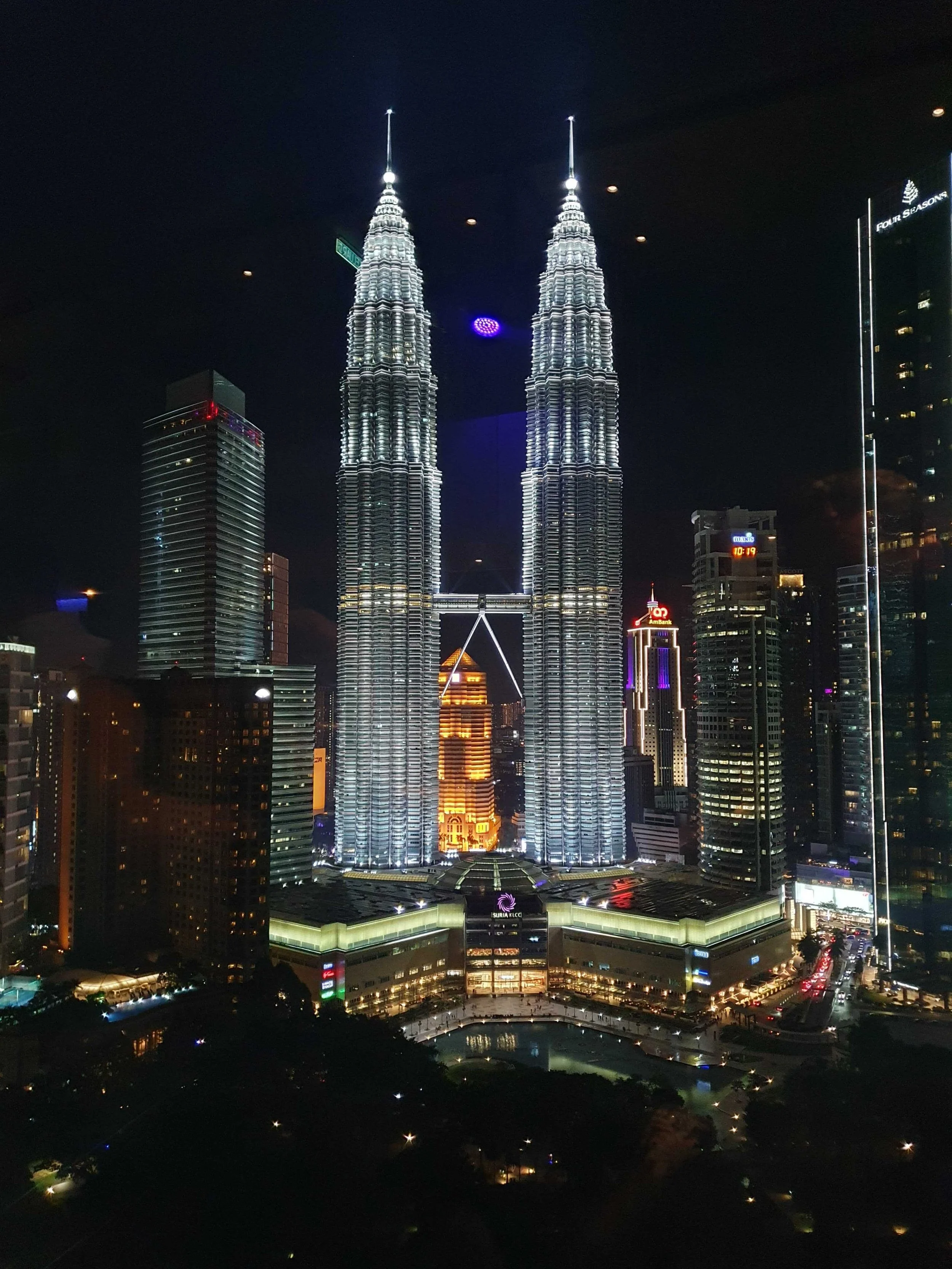 Nighttime view of the illuminated Petronas Twin Towers in Kuala Lumpur, Malaysia, with surrounding buildings and city lights.