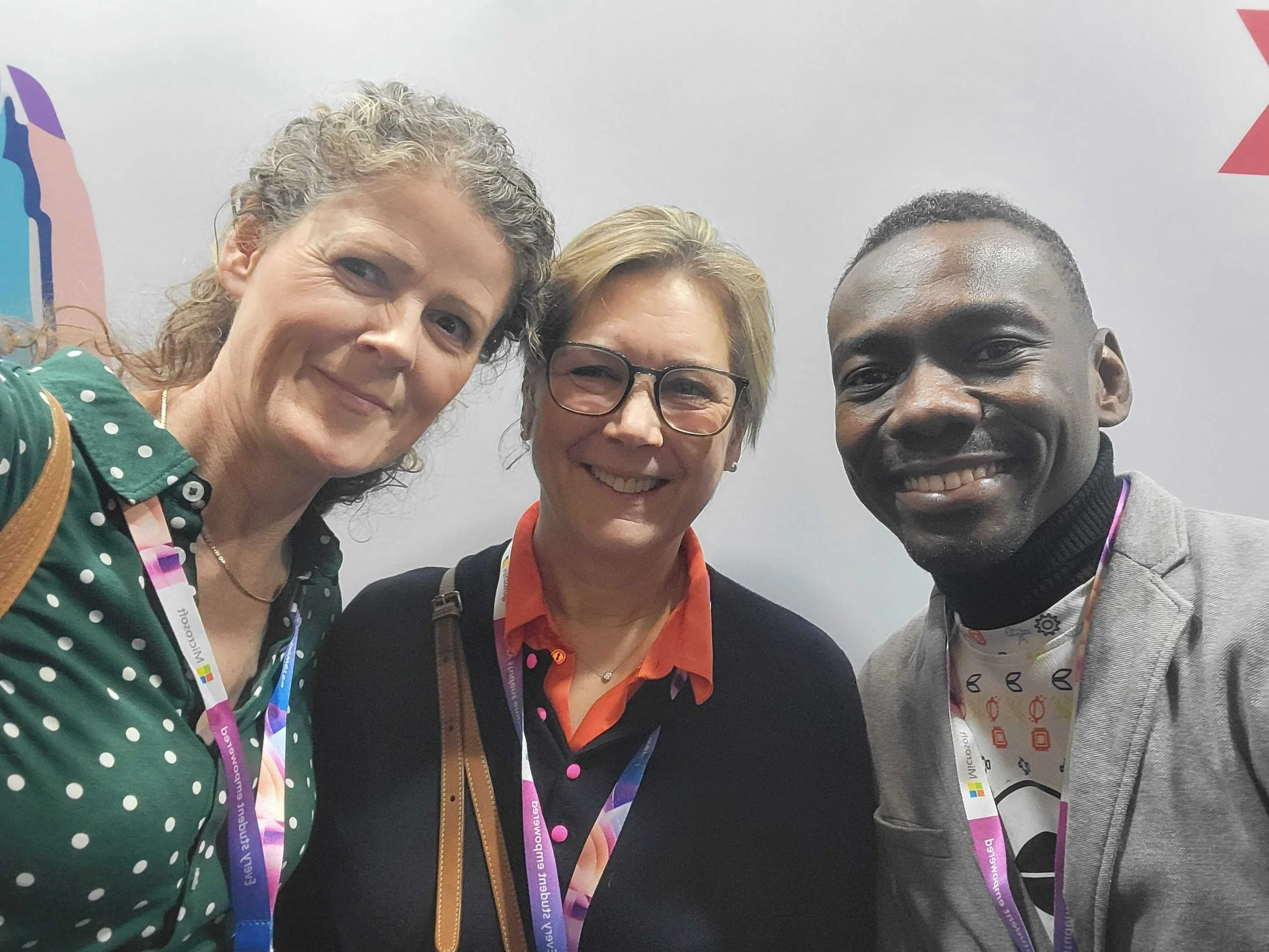 Three smiling people taking a selfie together at a conference or event, wearing conference badges and colorful clothing, with a plain background.