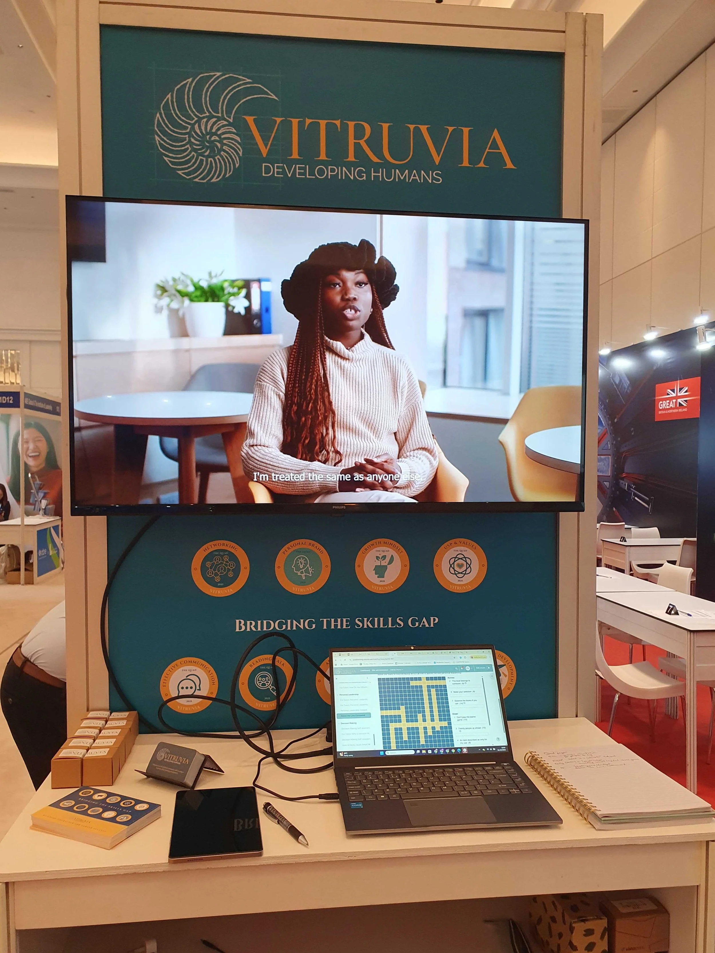 A booth display for Vitruvia with a large monitor showing an interview with a woman, titled 'Bridging the Skills Gap.' The table has informational brochures, a laptop, and a notebook, with some small boxed items and writing supplies. The booth backdr