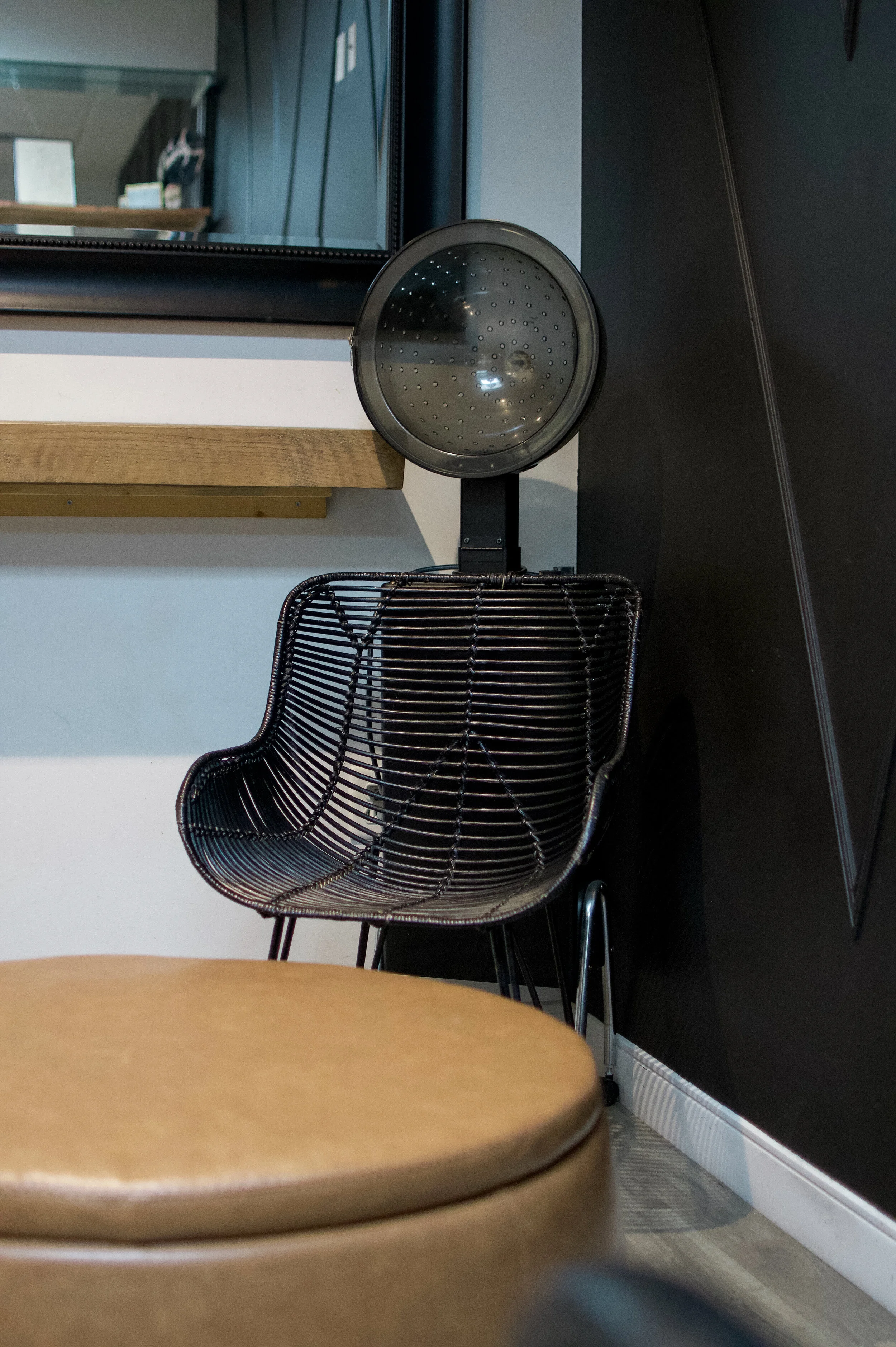 The image shows a black wire chair with a round wooden table in the foreground, a black sink spray nozzle and tiles on the wall in the background, and a mirror hanging above a wooden shelf.