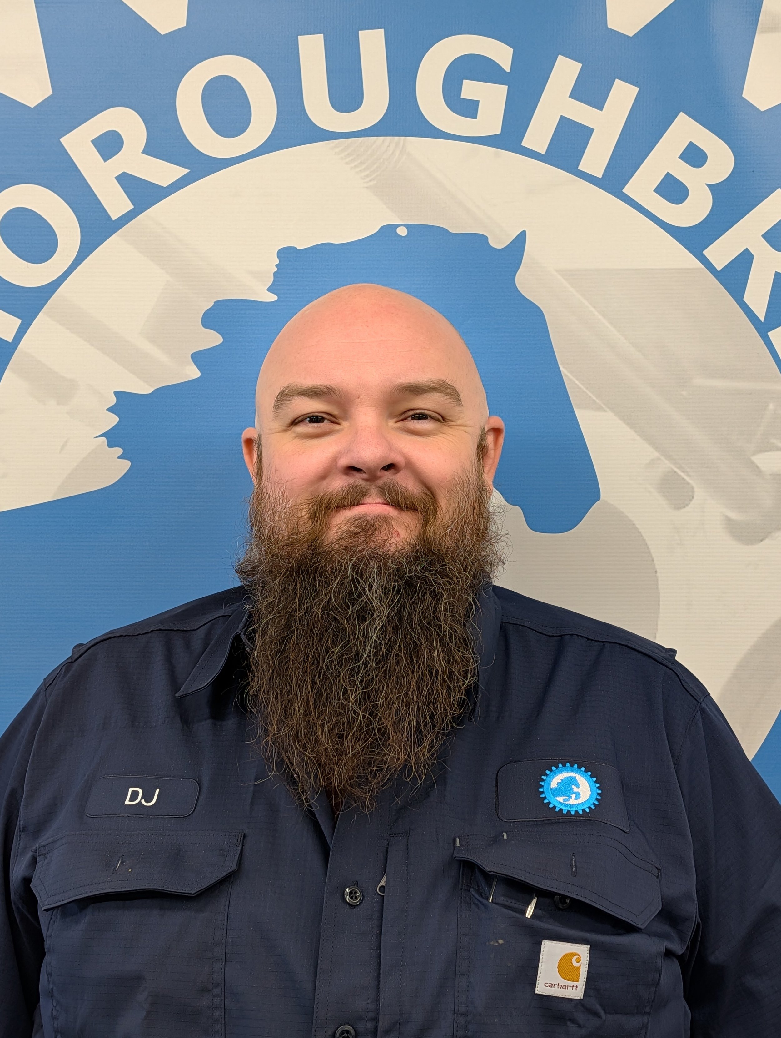 A man with a full beard and no hair smiling in front of a large sign that reads 'THOROUGHBRED' with a blue background and a silhouette of a horse.
