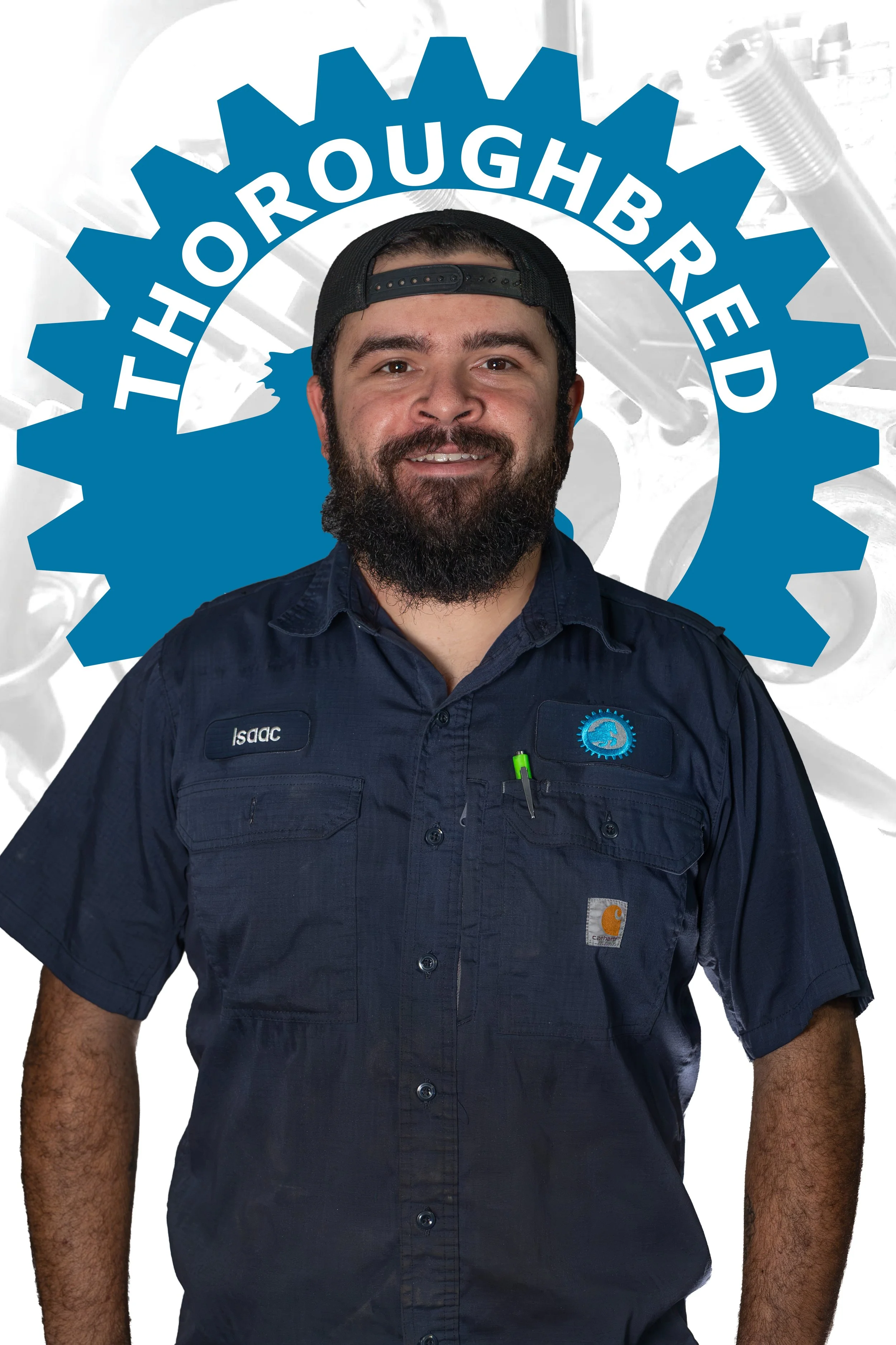A man with a beard and mustache, wearing a navy blue Carhartt work shirt with a name tag that says Isaac and a logo patch, smiling and standing in front of a background with a large blue gear and the words "Thoroughbred Motors" written across it.