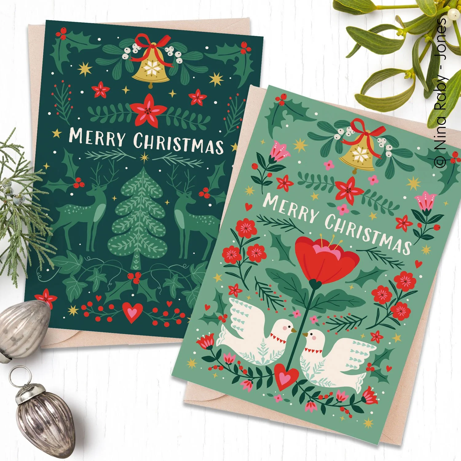  Folk inspired 2 pack festive holiday designs and message "Merry Christmas". One card features reindeer and a Christmas tree with holly and bells, the other has a red flower, two white doves, and holly. 