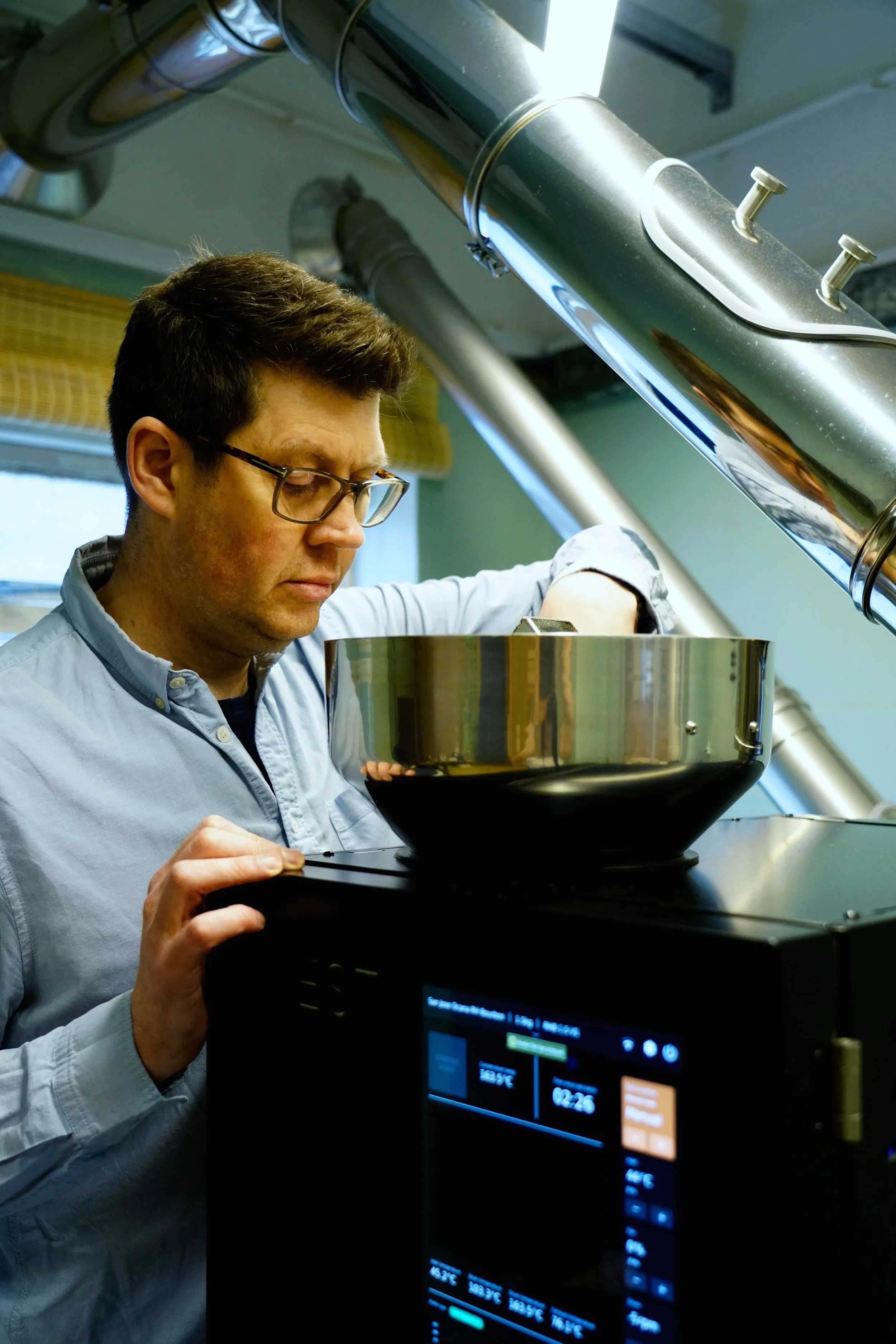 A coffee roasting man wearing glasses examines the hopper where the green beans are put. Industrial venting in the backdrop from the machine.
