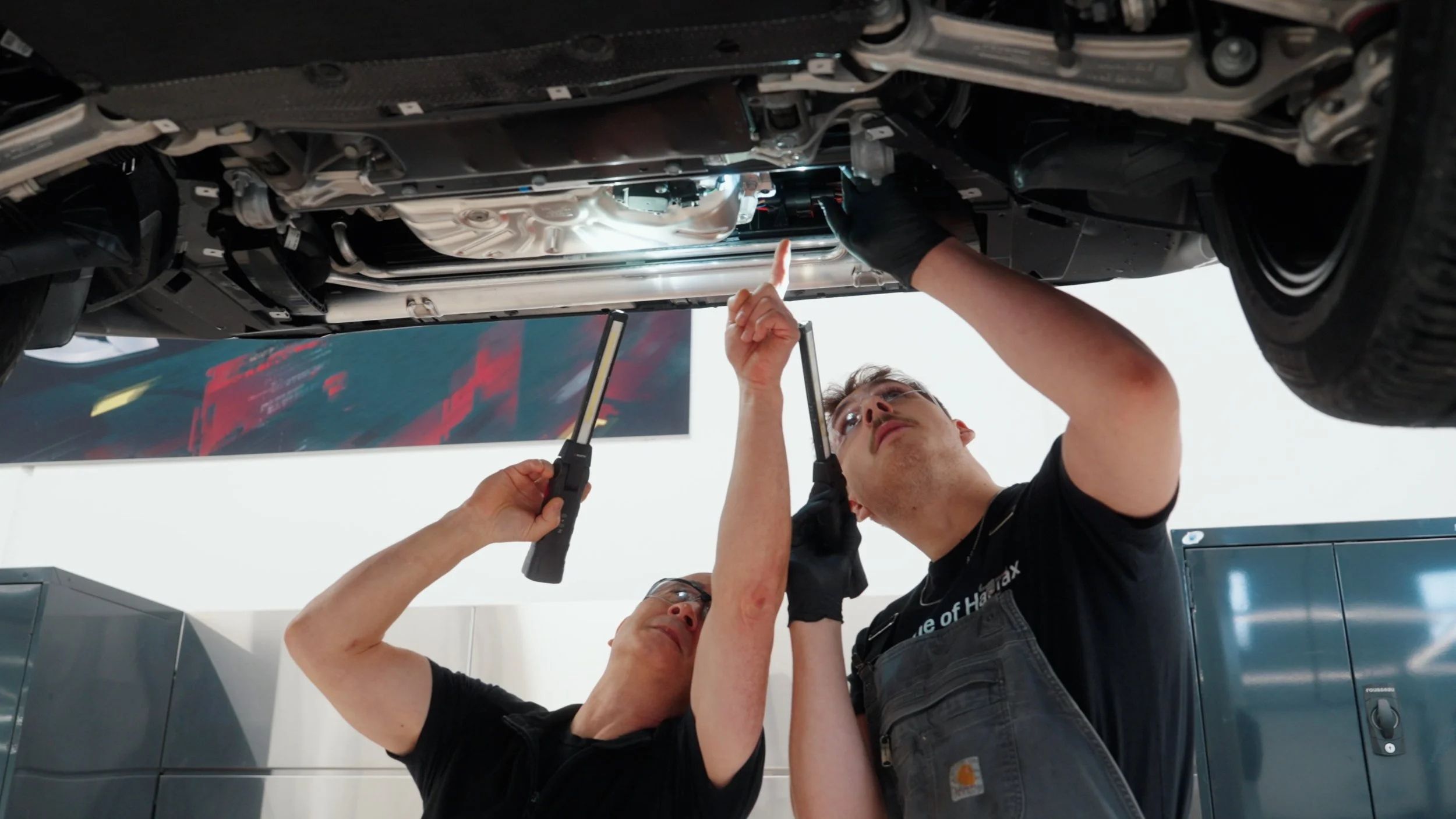 Two mechanics working underneath a car, inspecting and repairing components, with one mechanic pointing and the other holding tools.