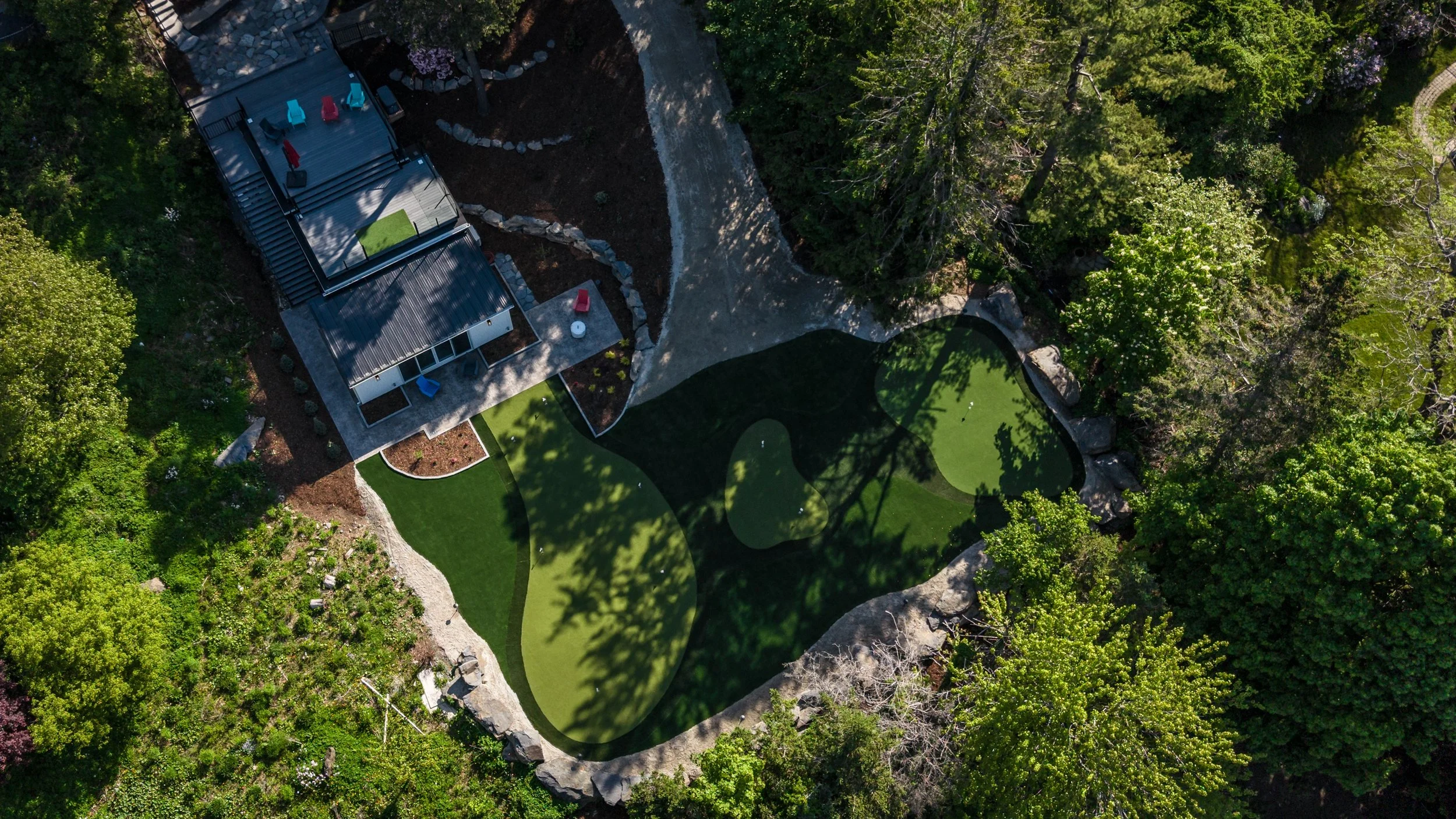 Aerial view of a backyard with a deck, a pond, a mini golf course, and surrounding trees
