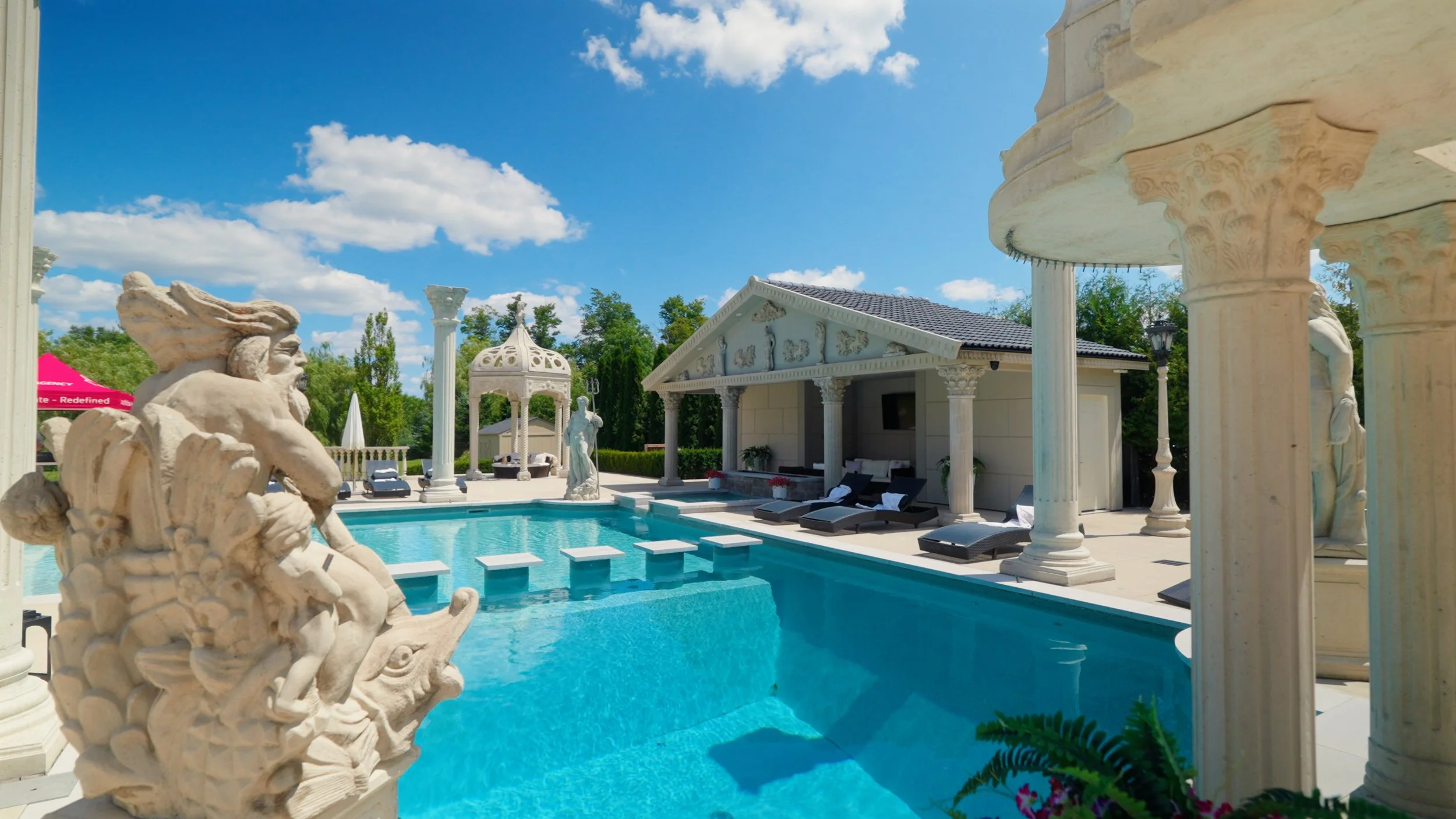 Luxury backyard with a swimming pool surrounded by classical Greek-style white sculptures, columns, and a small pavilion under a blue sky with scattered clouds.