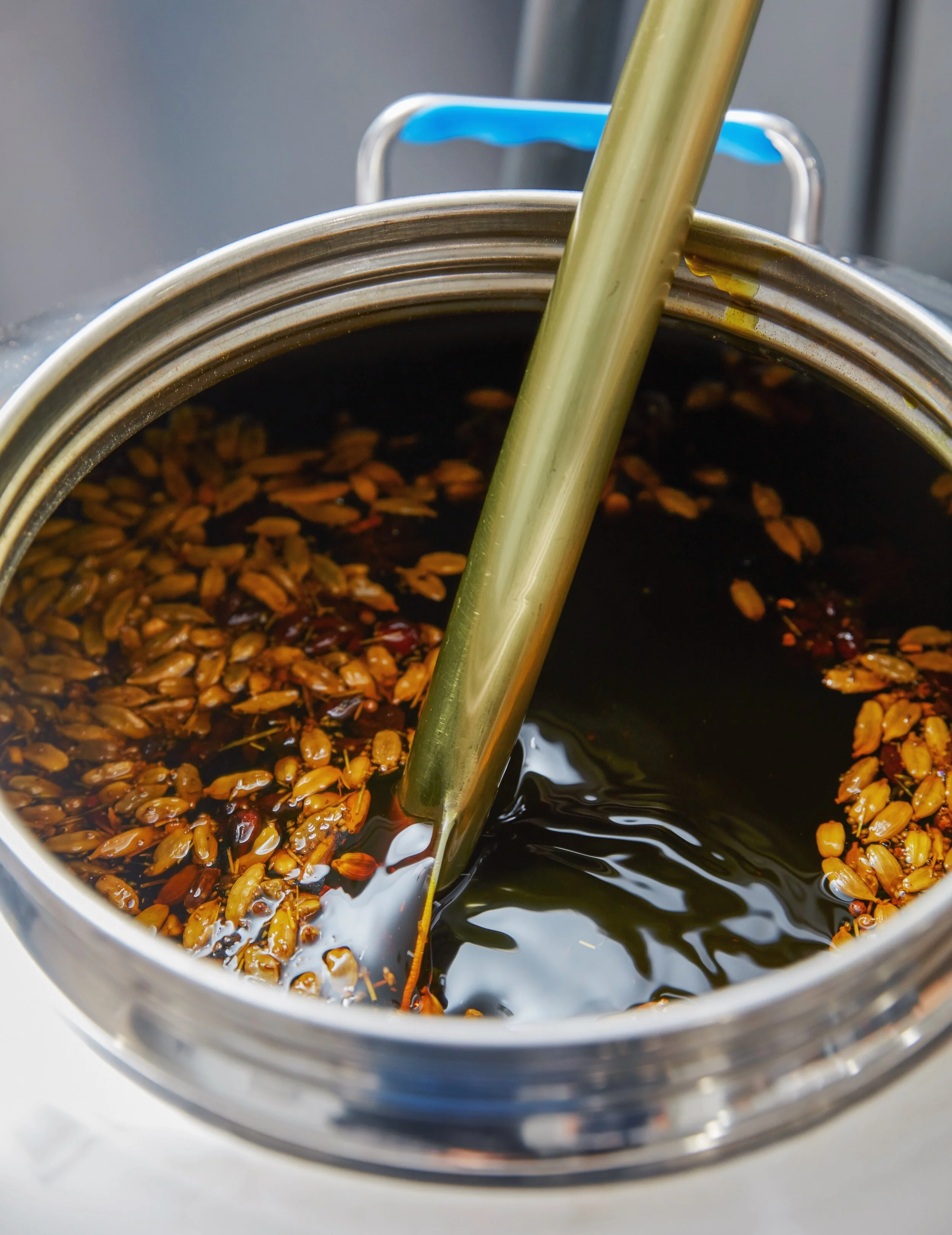 Close-up of a container filled with a dark liquid and fermented rice grains, with a stainless steel paddle stirring the mixture.