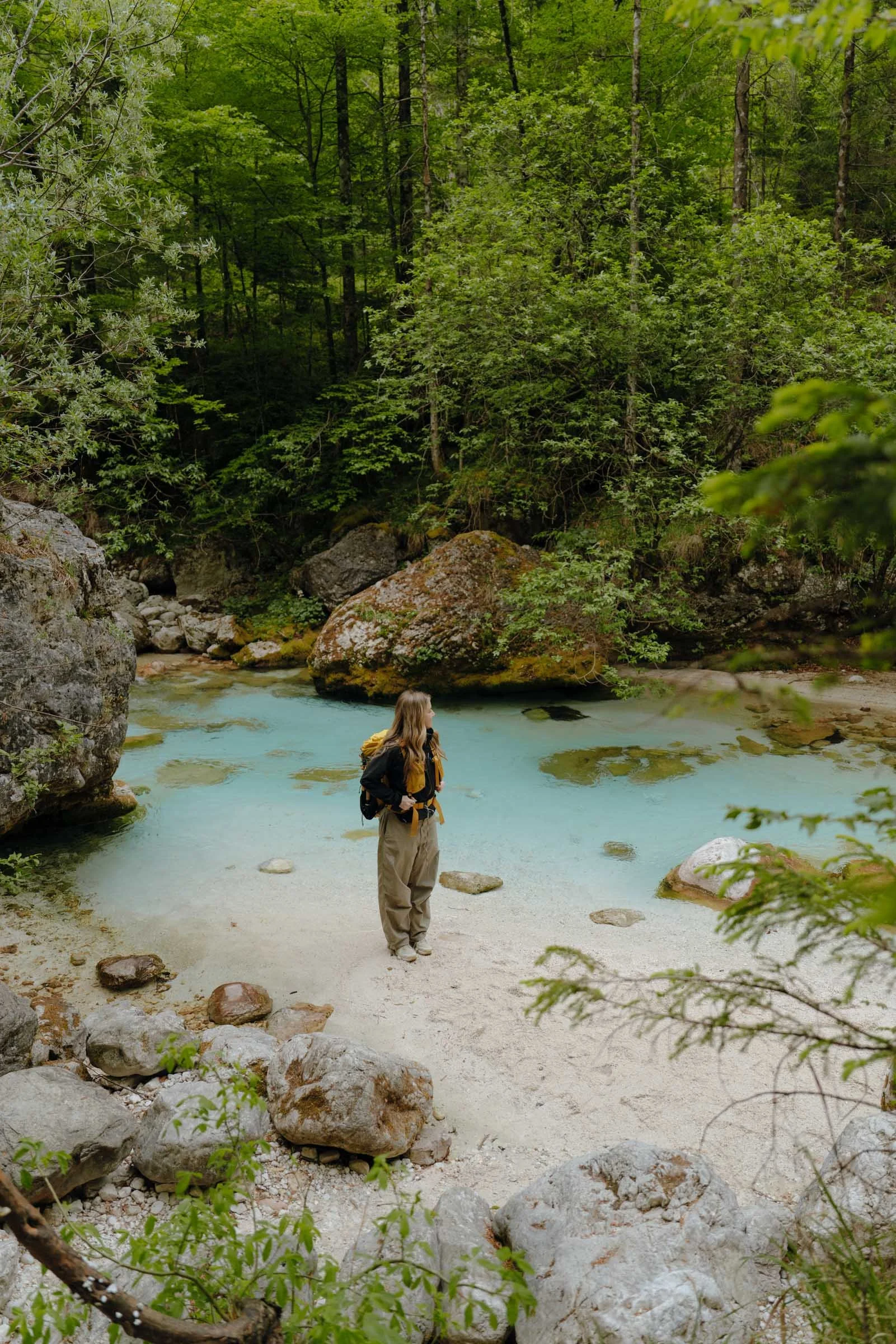 Woman standing in hiking gear next to a bright blue river in the Soca Valley, Alpe Adria Trail, Slovenia