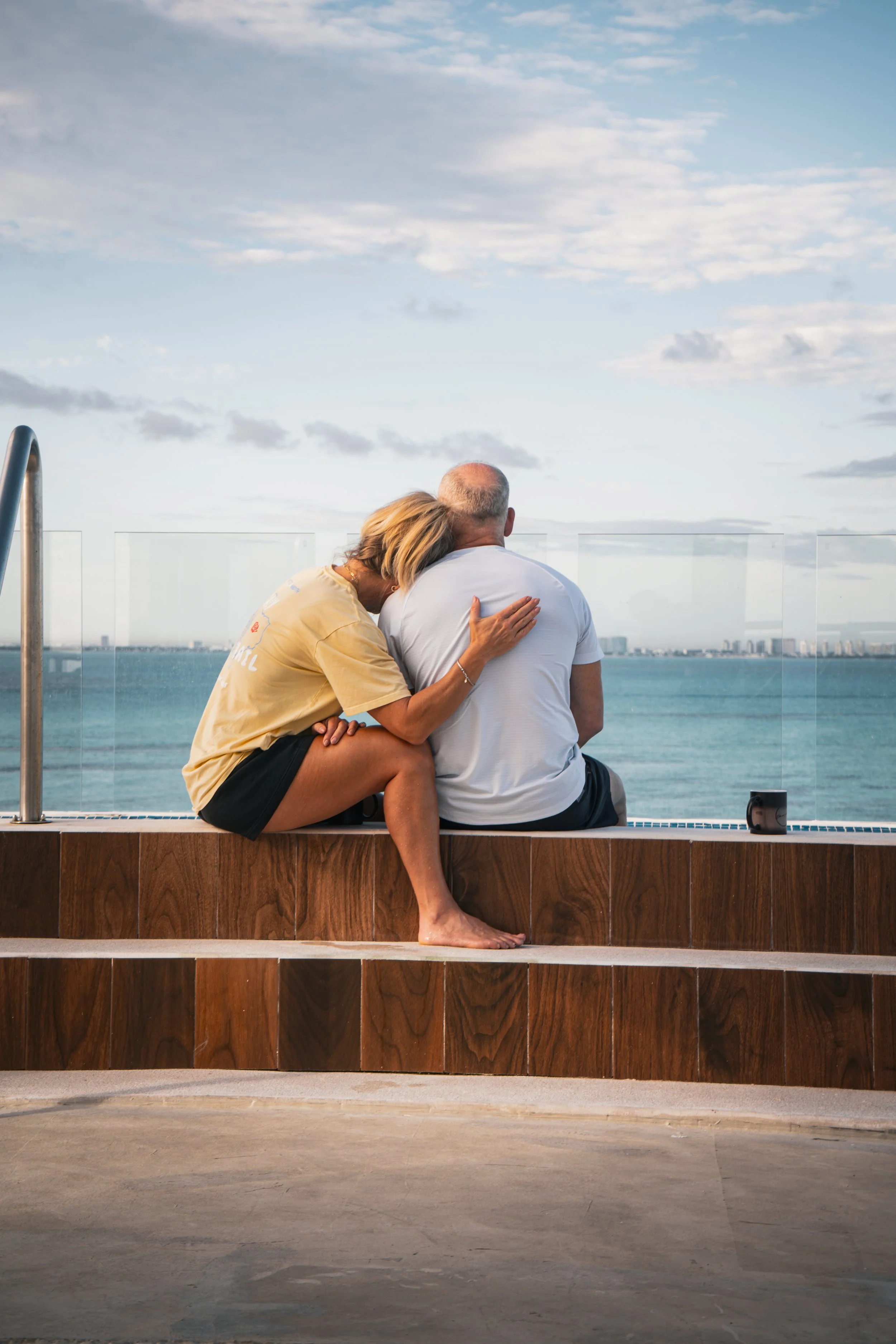 An elderly couple sitting on a ledge overlooking the ocean, with the woman resting her head on the man's shoulder at sunset.