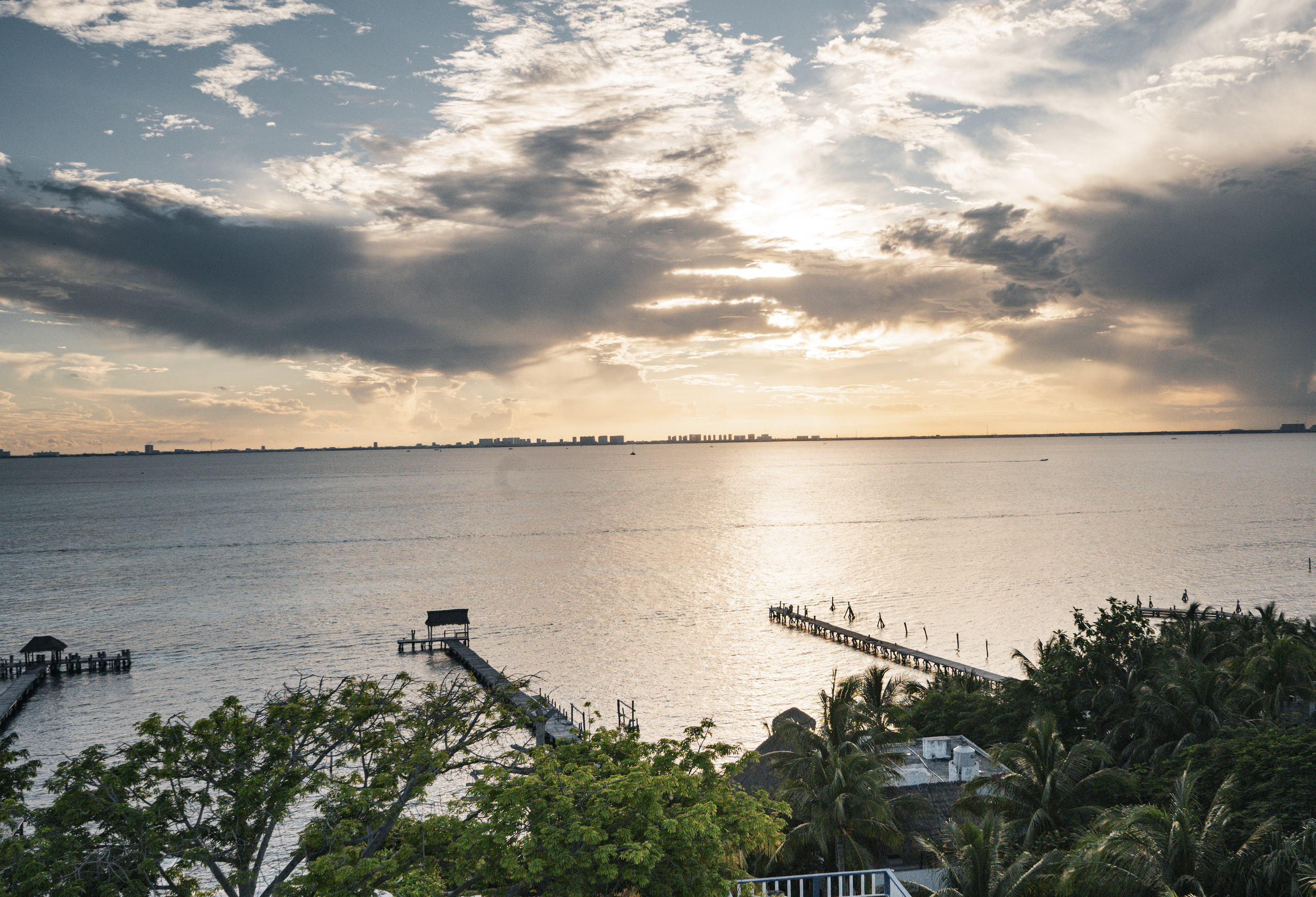 Sunset over a body of water with two wooden docks extending into the ocean, surrounded by green trees and palm trees in the foreground, and a city skyline far in the distance.