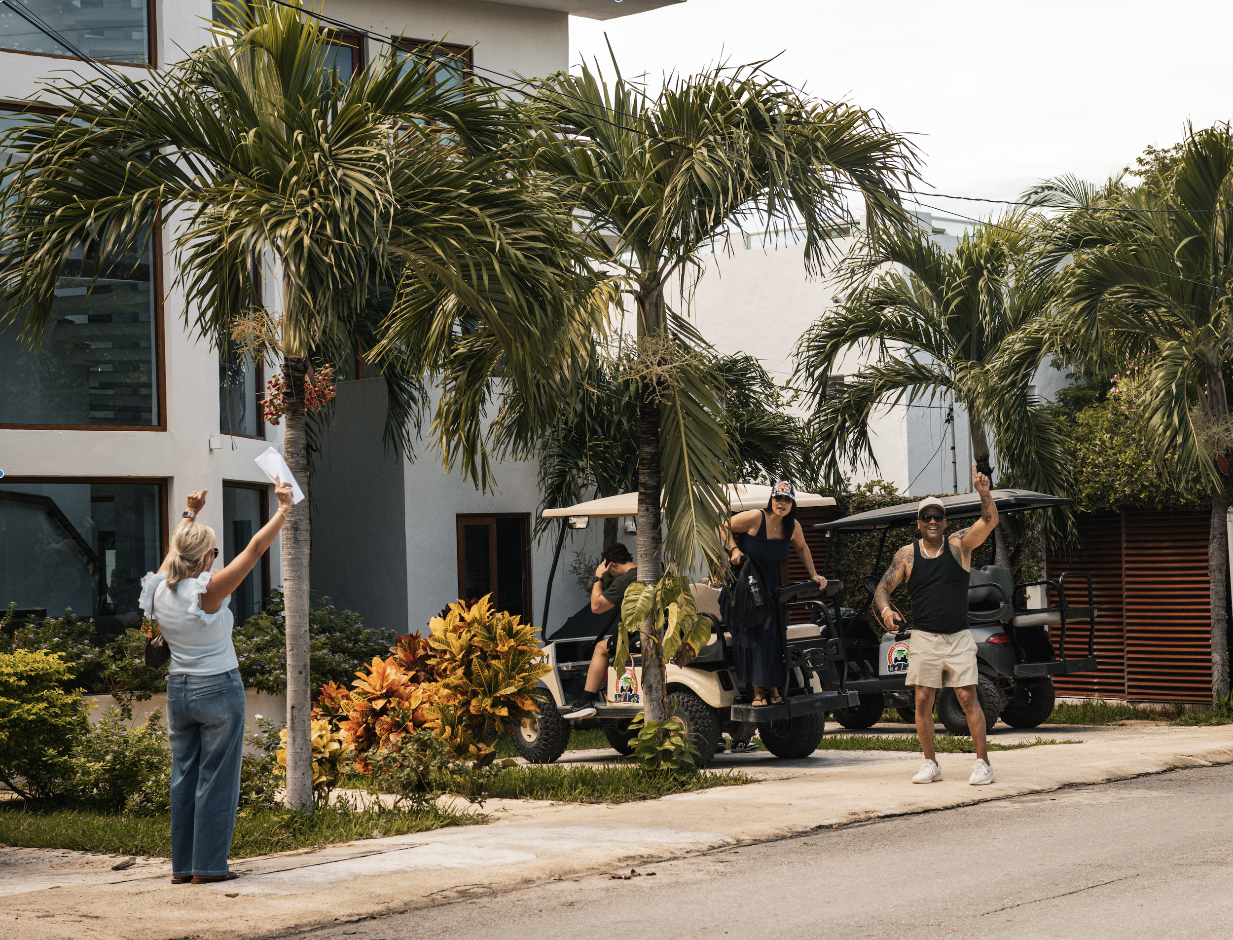 A woman standing on a sidewalk taking a photo of two people riding in golf carts, surrounded by palm trees and tropical plants outside a modern building.