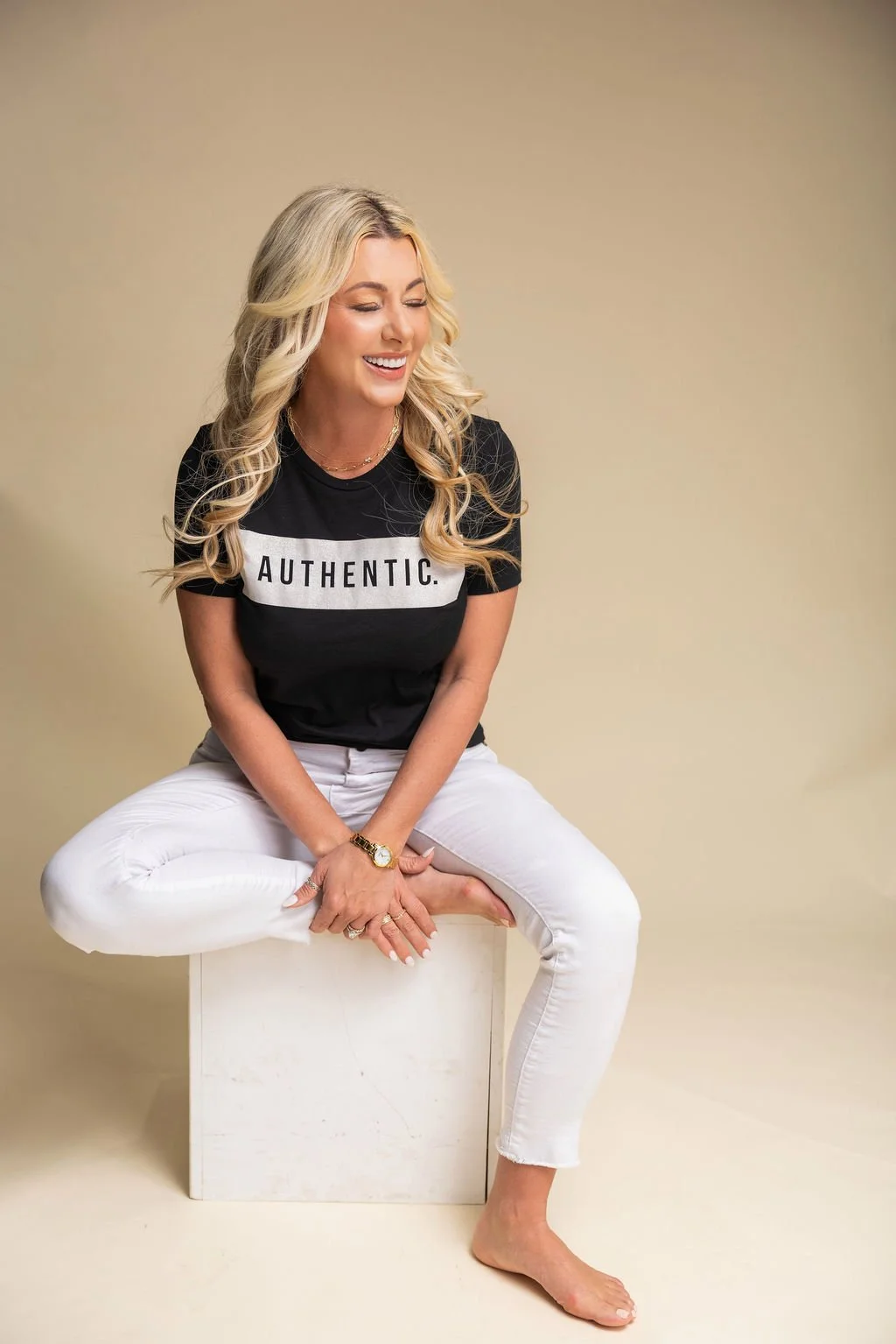 A woman with long blonde hair sitting on a white box, wearing a black T-shirt with the word 'AUTHENTIC.' printed on it, and white pants. She is smiling and looking to her right, with a beige background.