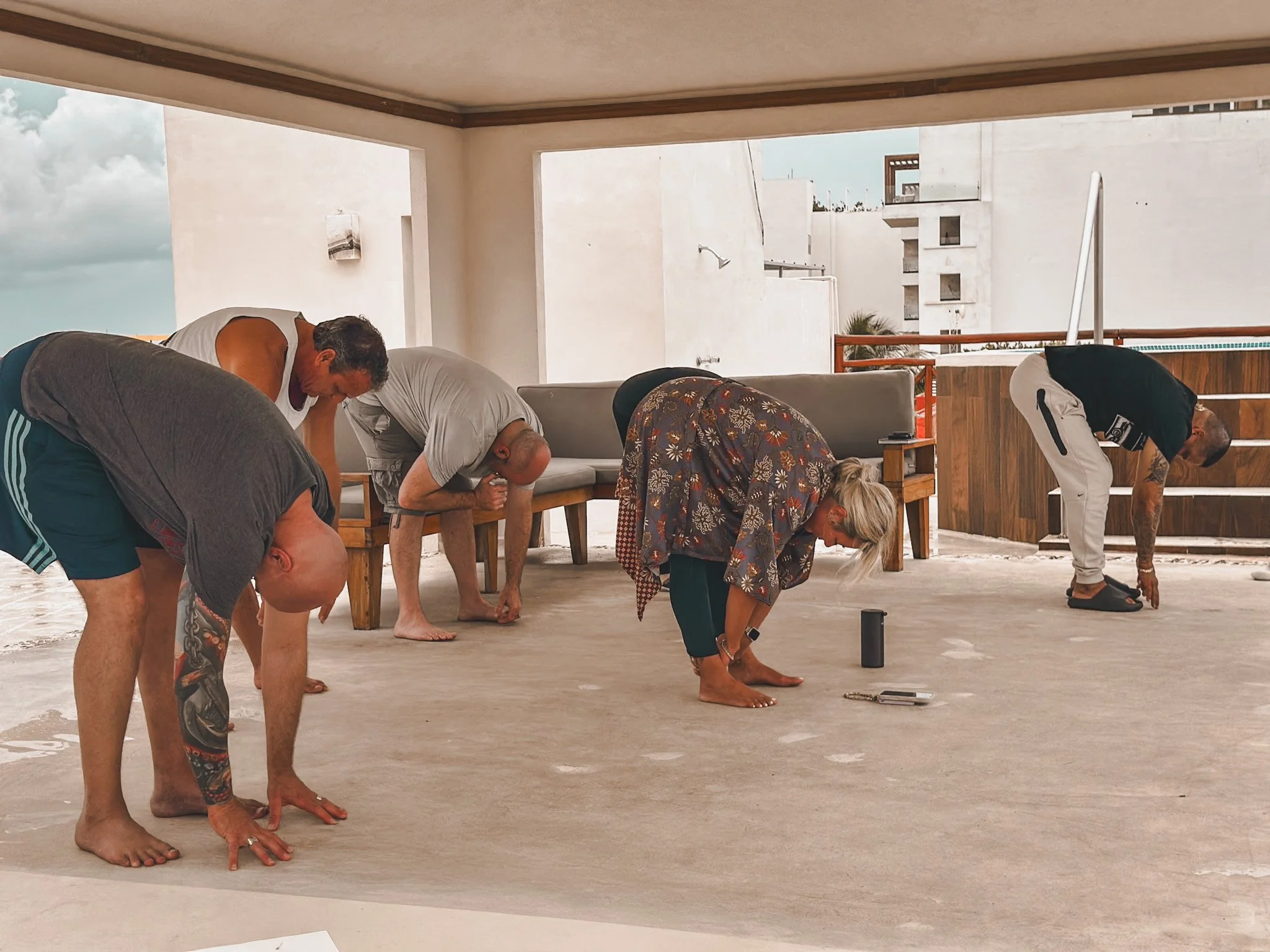 Five people practicing yoga or stretching indoors on a concrete floor, bending forward with hands on the ground.