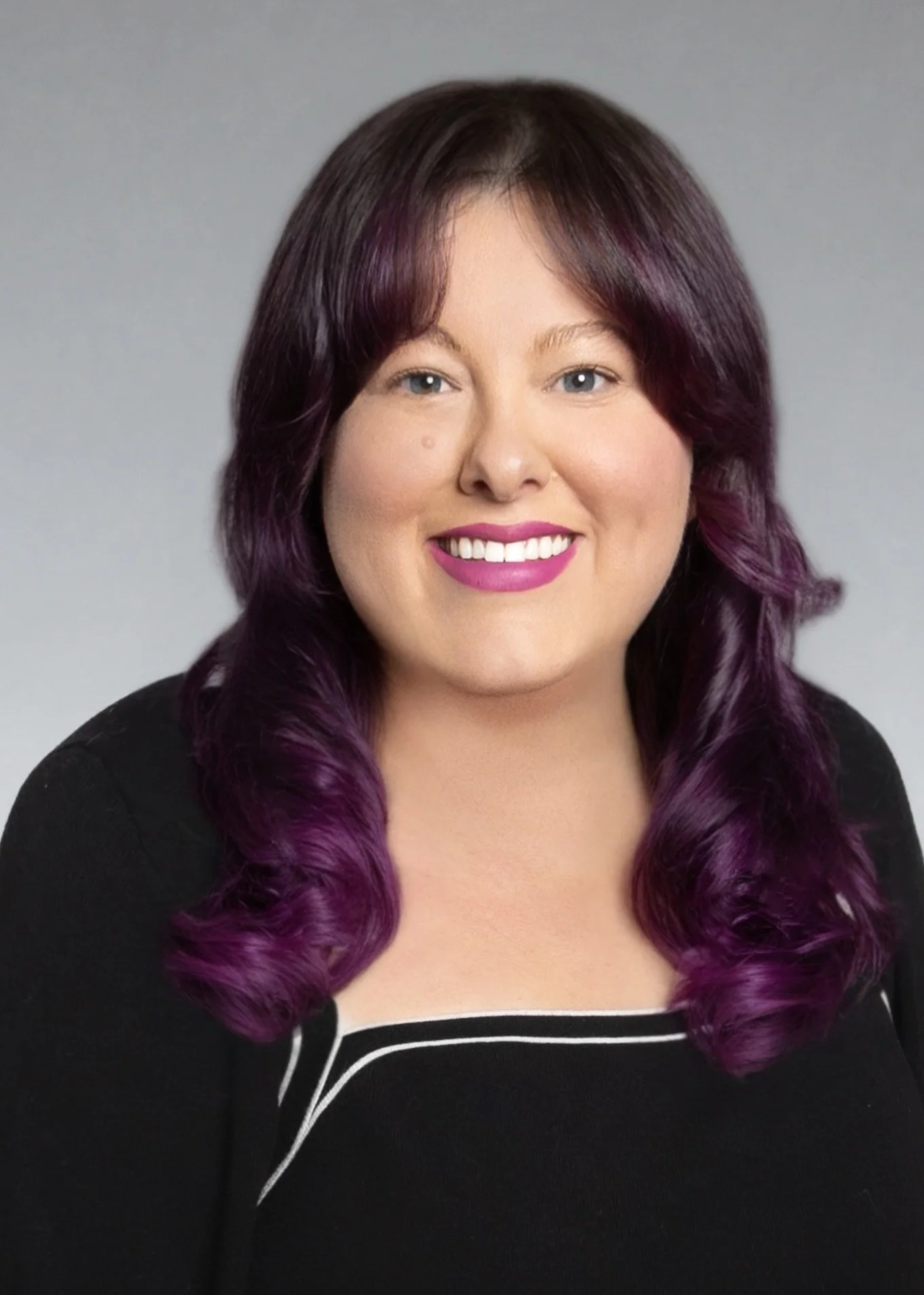 Hillary Crow, SLP, speech therapist, a woman with shoulder-length purple hair, blue eyes, and pink lipstick smiling at the camera, wearing a black top with white trim and a plain gray background, providing speech therapy near you in Orange County.