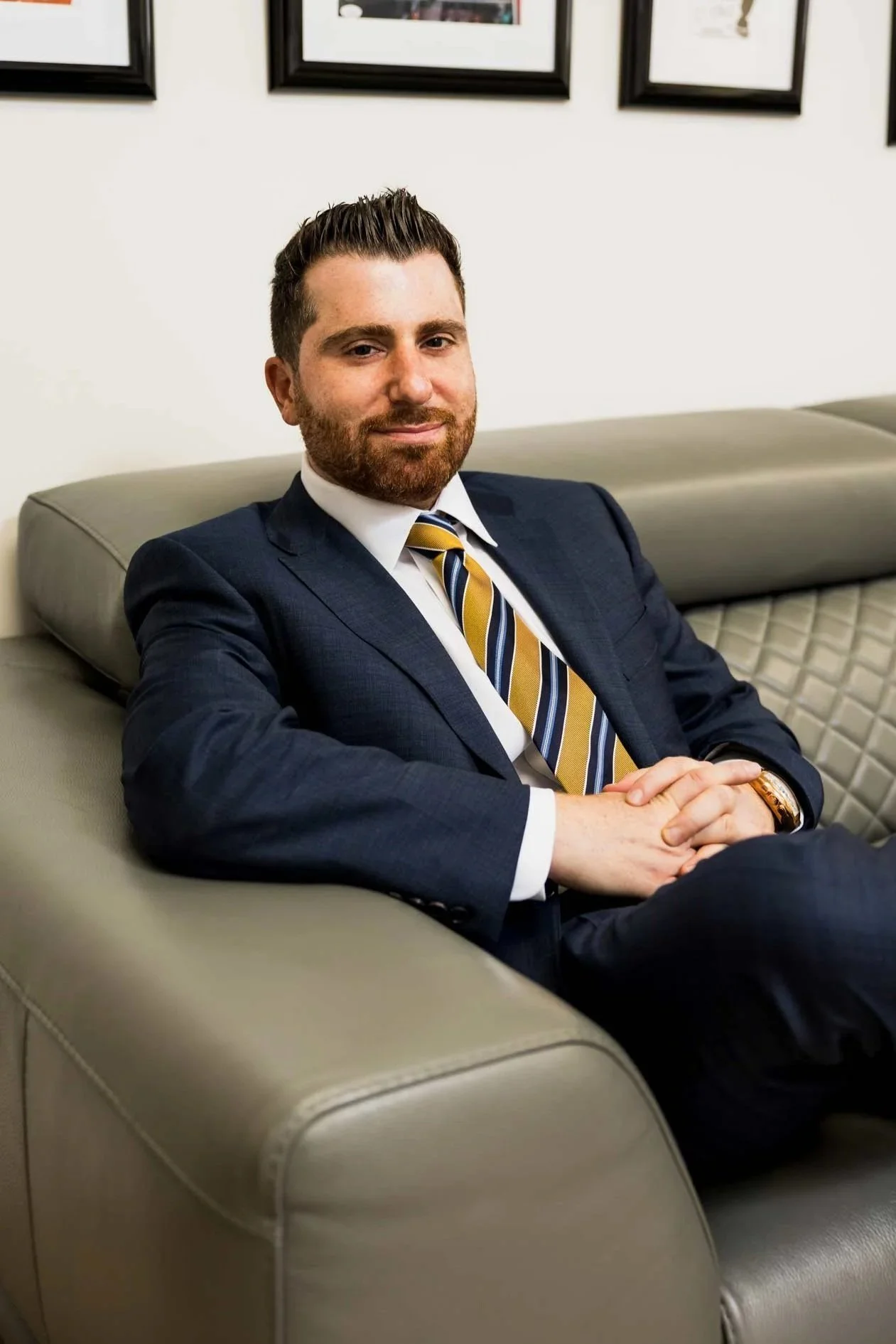 A man in a dark blue suit with a striped yellow and blue tie, sitting on a beige leather sofa with his hands clasped, in a professional office setting with framed pictures on the wall behind him.