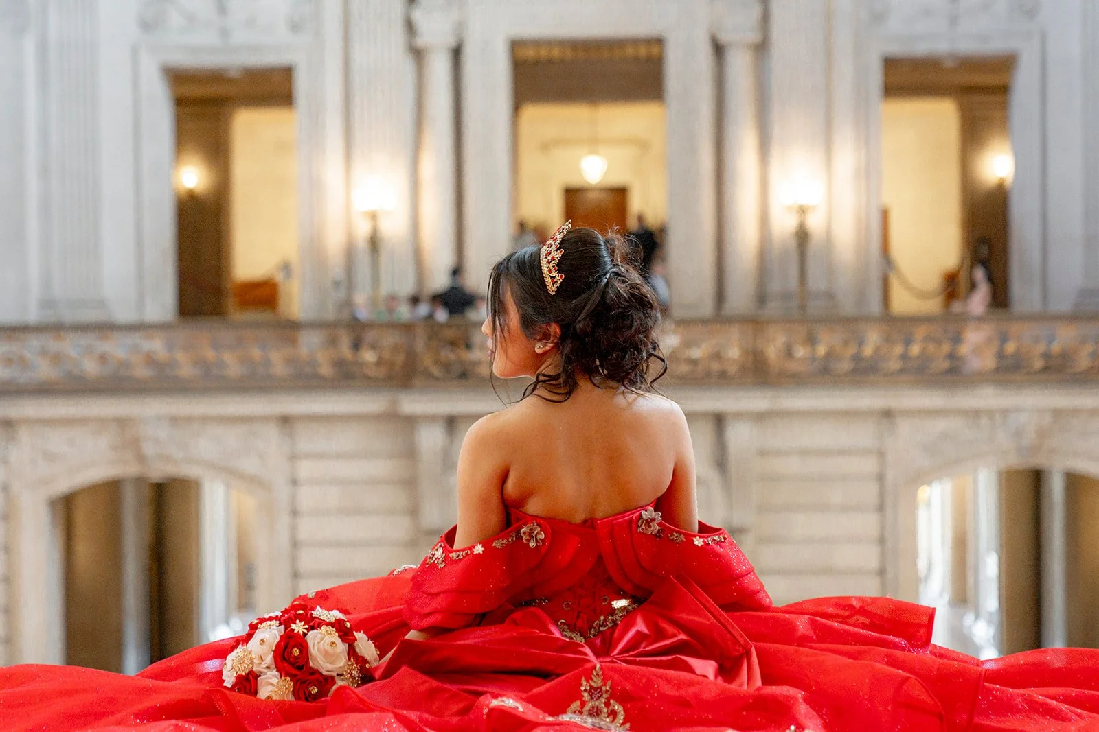 A woman in a red wedding dress sitting on a staircase in a grand hall, facing away from the camera, with a bouquet of flowers beside her.