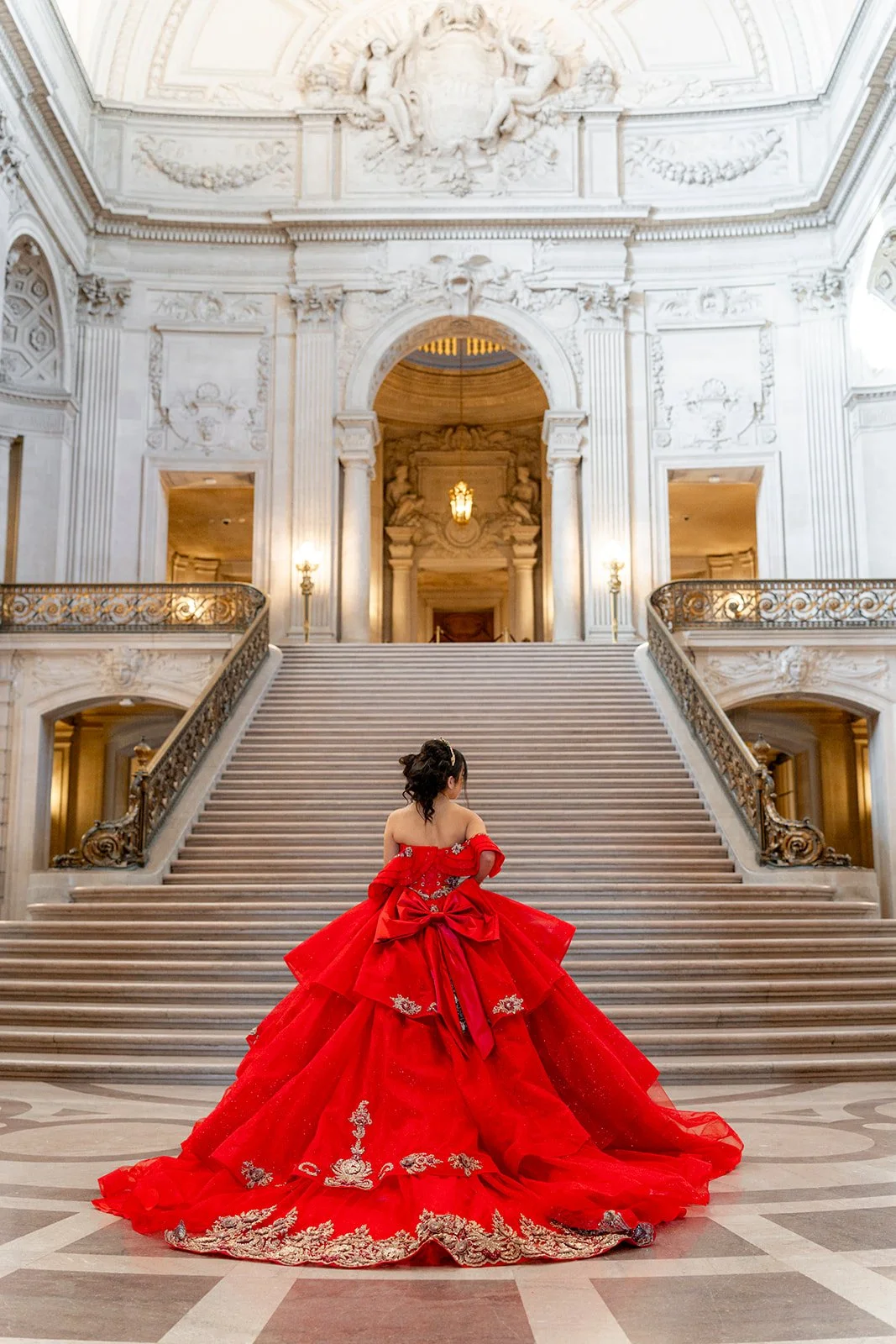 A woman in a red, intricately designed ball gown stands at the bottom of a grand staircase inside a luxurious building with ornate white interior and high ceilings.