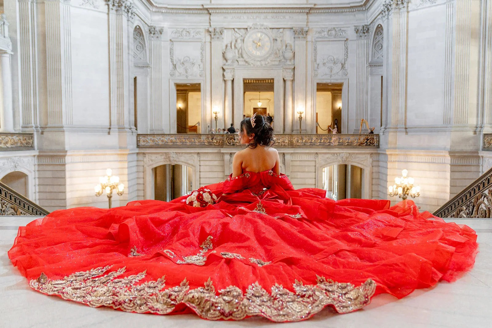 A woman in a red wedding dress with intricate gold embroidery sitting on a marble staircase inside a grand, historic building with ornate architecture and chandeliers.