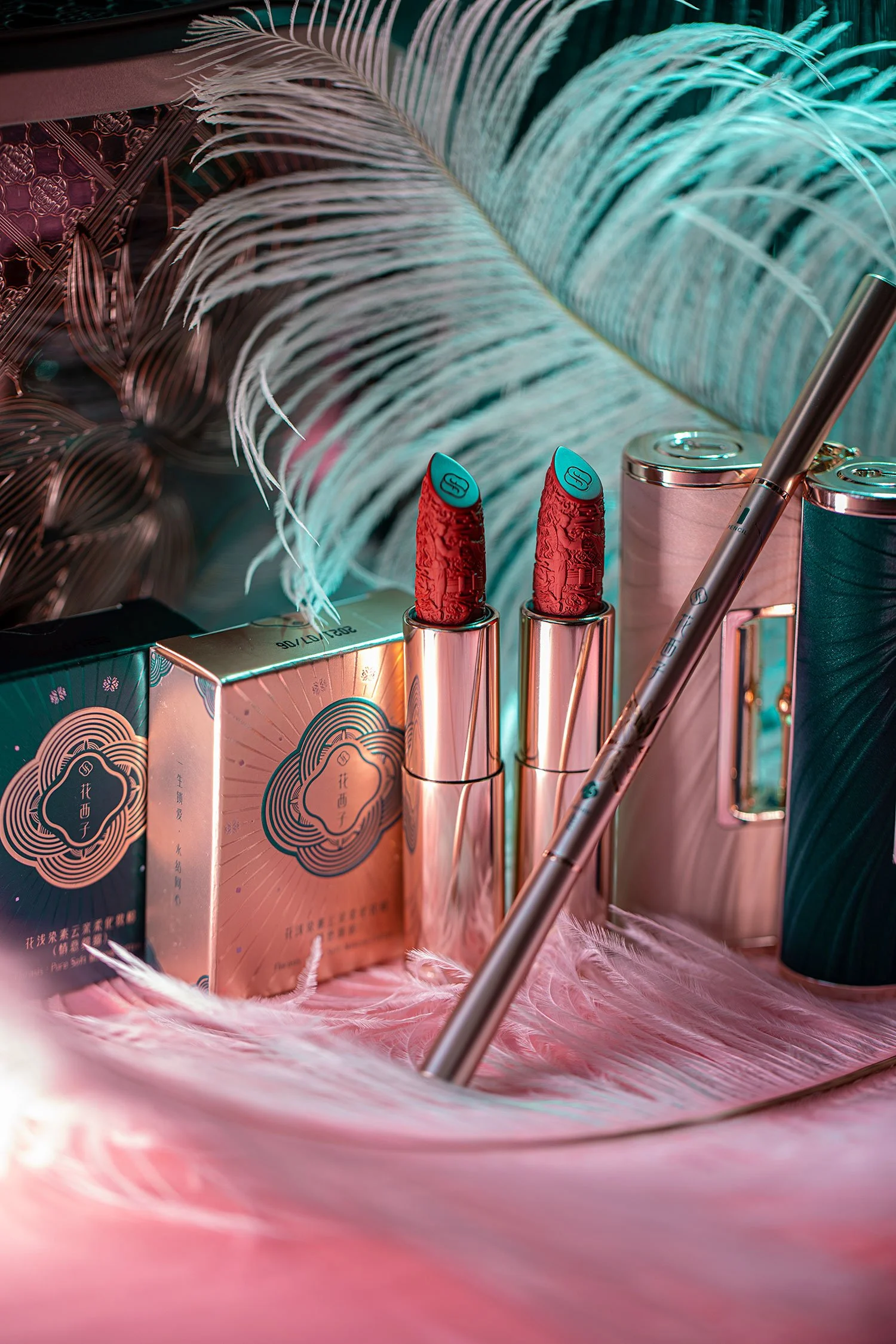 Two tubes of red lipstick with blue tips, surrounded by packaged cosmetics, a metallic pen, and pink and teal cylindrical containers on pink feathers, with large teal feathers in the background.