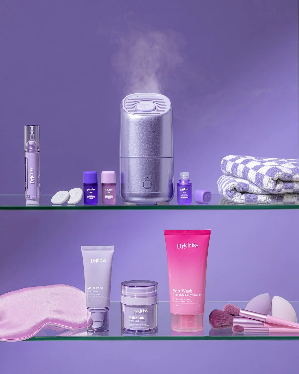 Purple-themed skincare display with Dr.Idriss products, facial steamer, folded towels, face mask, cleanser, moisturizer, makeup brushes, and spa accessories.