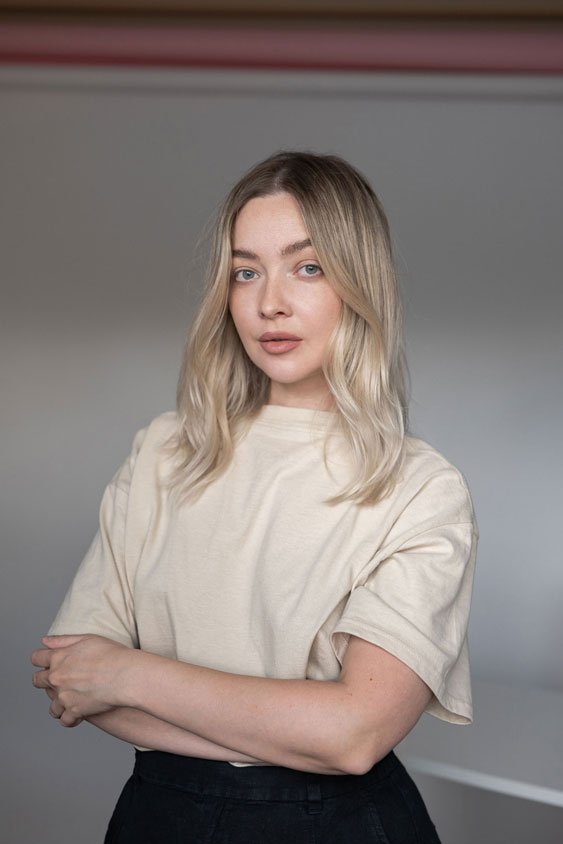 Blonde woman in a beige oversized T-shirt and black pants posing with crossed arms against a gray background