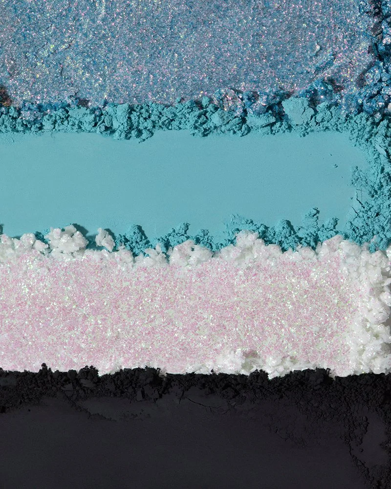 Close-up of four different types of colorful, shimmery powders or pigments arranged in horizontal layers, including glittery purple, bright blue, pastel pink, and black.