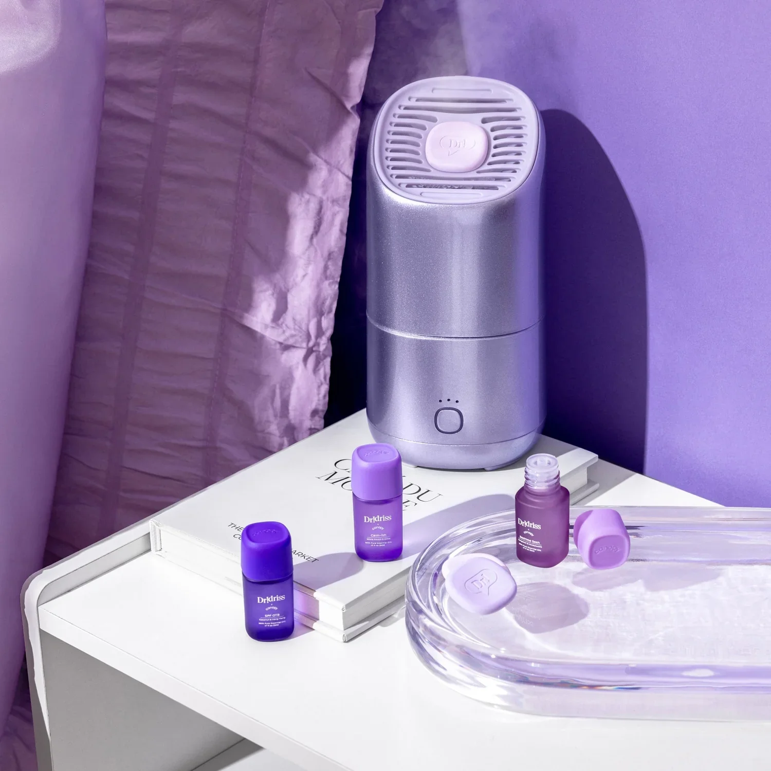 Purple Dr Idriss skincare bottles and a silver air purifier on a white bedside table beside books and a glass tray.
