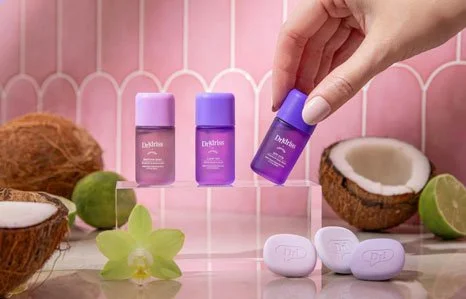 Hand holding a purple Dr. Idriss+ skincare bottle beside matching products, coconuts, limes, and an orchid on a pink background