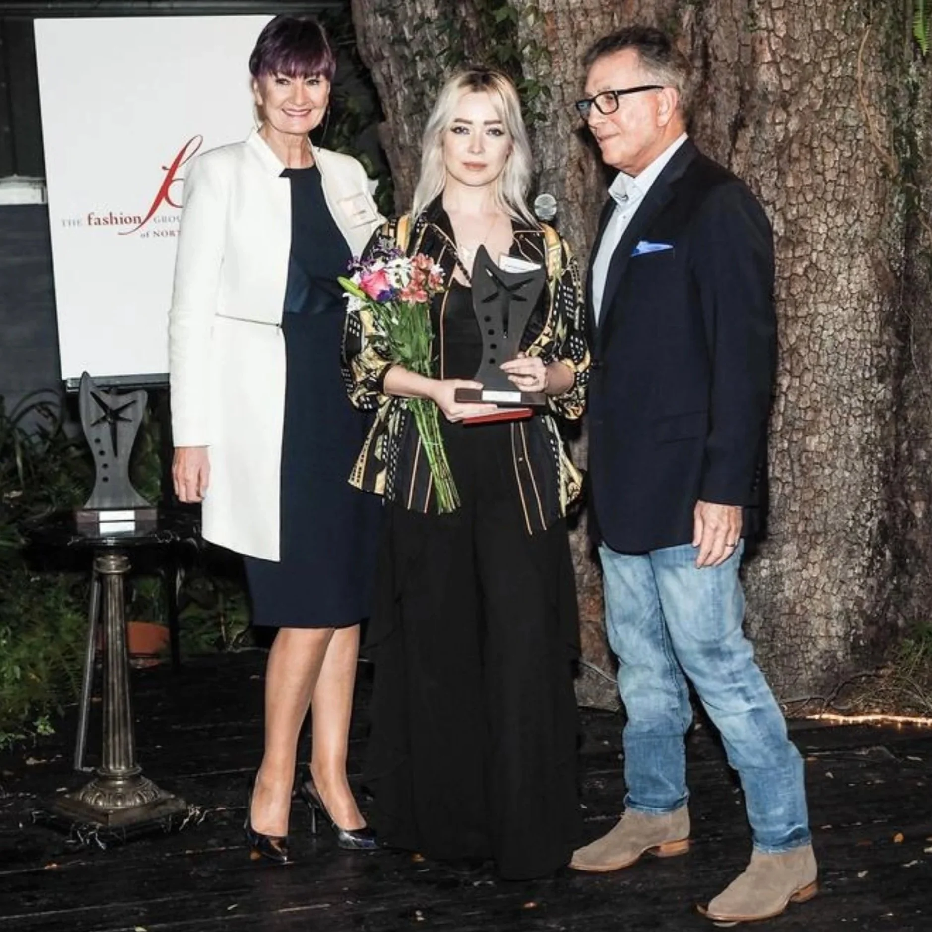 Three people pose at a fashion awards event, with a blonde woman holding flowers and a trophy between two presenters.