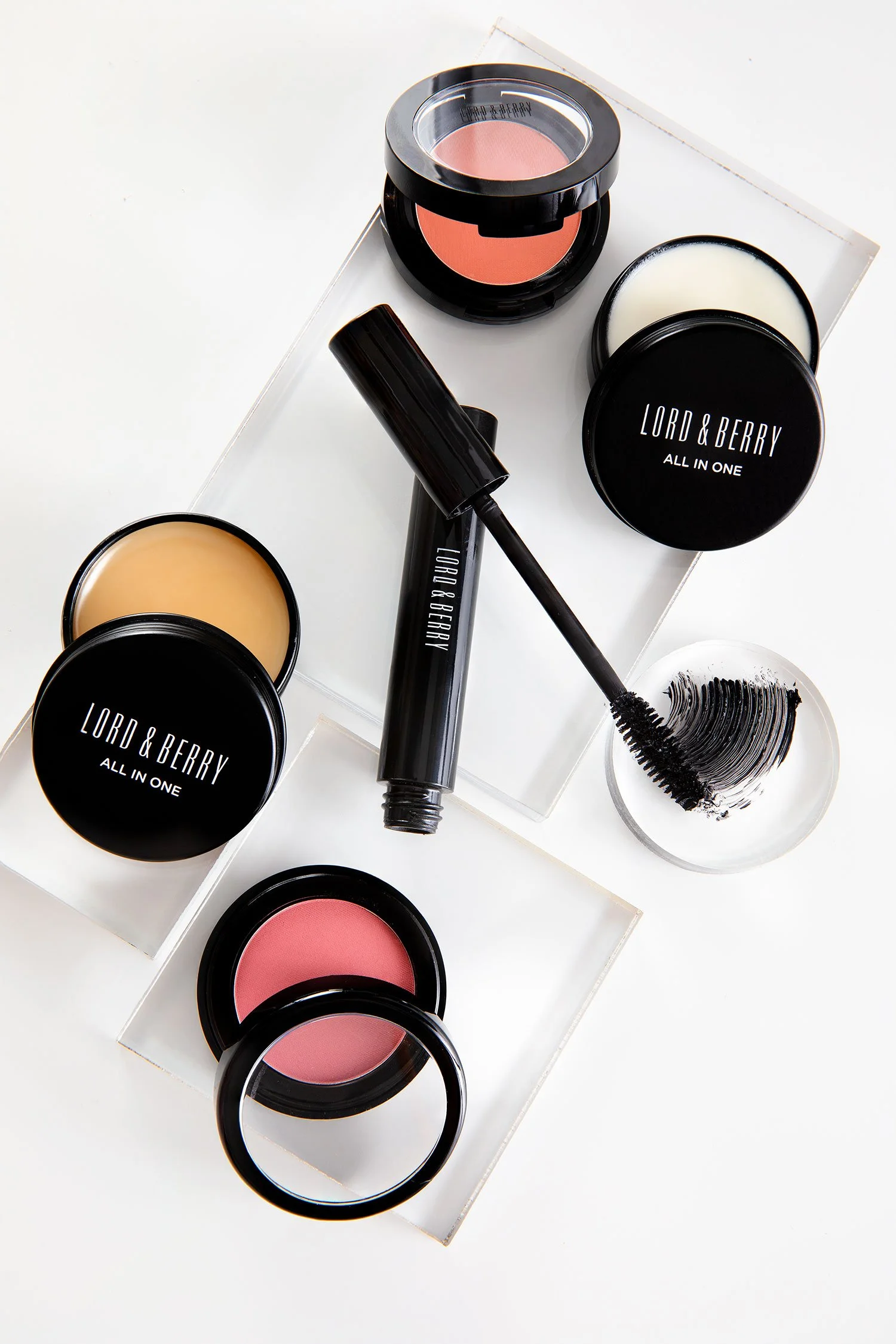 Makeup products including blush, foundation, mascara, and lip gloss on a white surface with clear display trays.