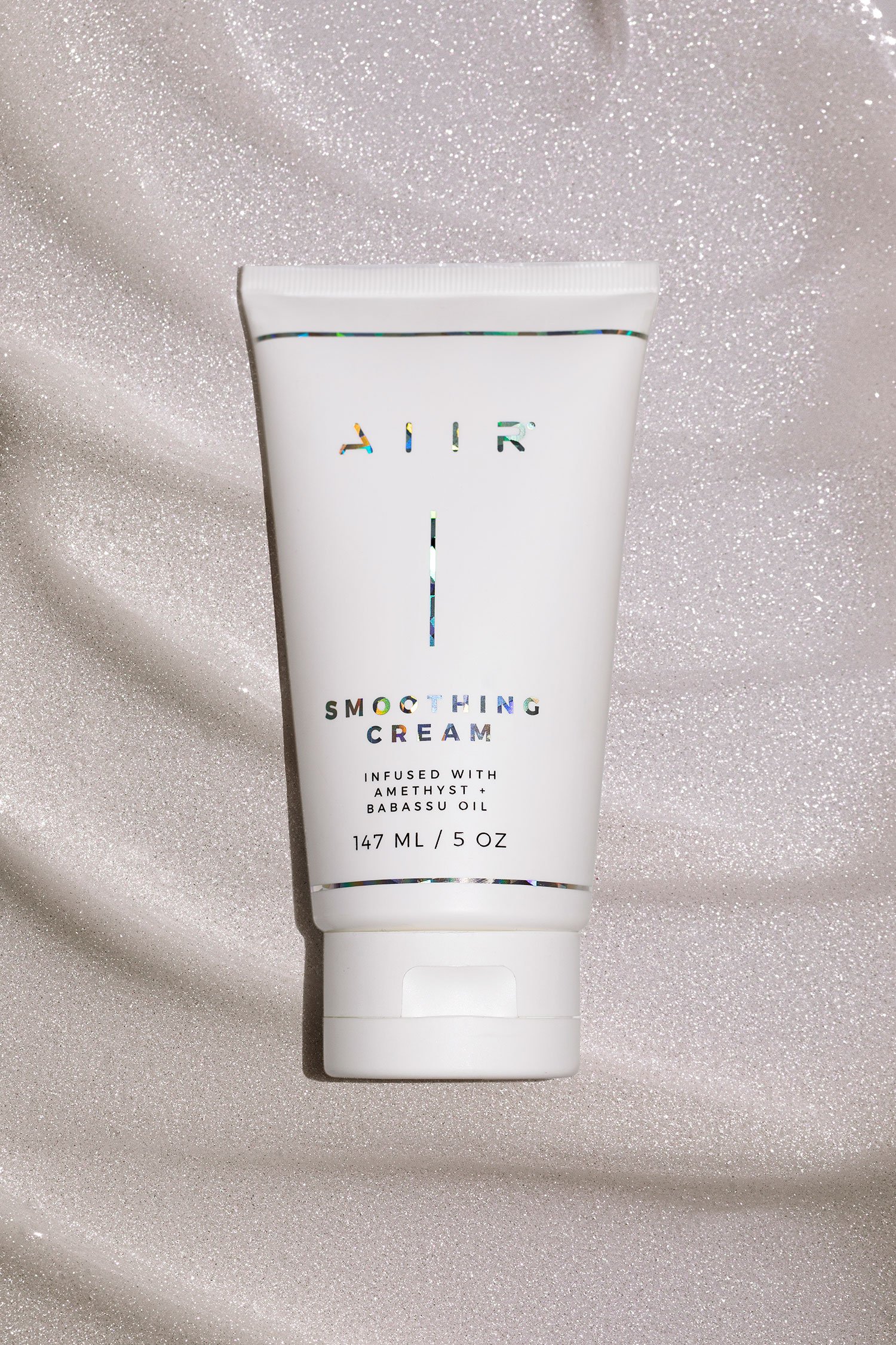A white tube of AER Smoothing Cream with holographic lettering, placed on a shimmery silver fabric background.