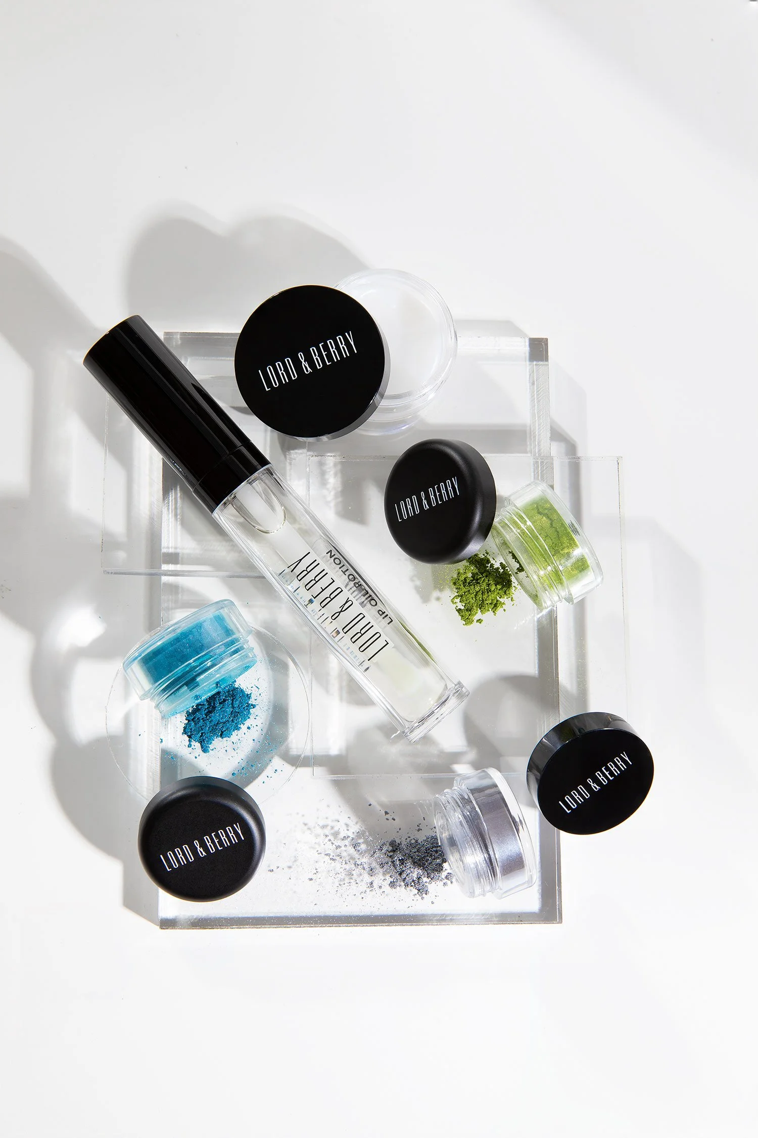 Beauty products including jars of colorful powder, a rollerball fragrance, and containers labeled "Lord & Berry" on a clear acrylic tray.