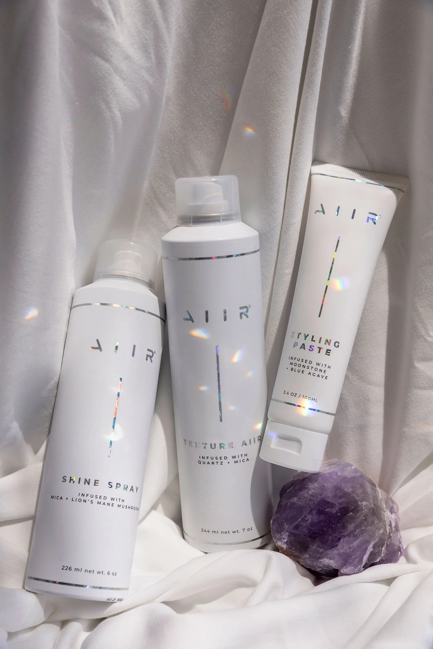 Three bottles of hair styling products placed on white fabric with a purple crystal gemstone nearby. The bottles are white with holographic accents and labels indicating shine spray, texture air, and styling paste.