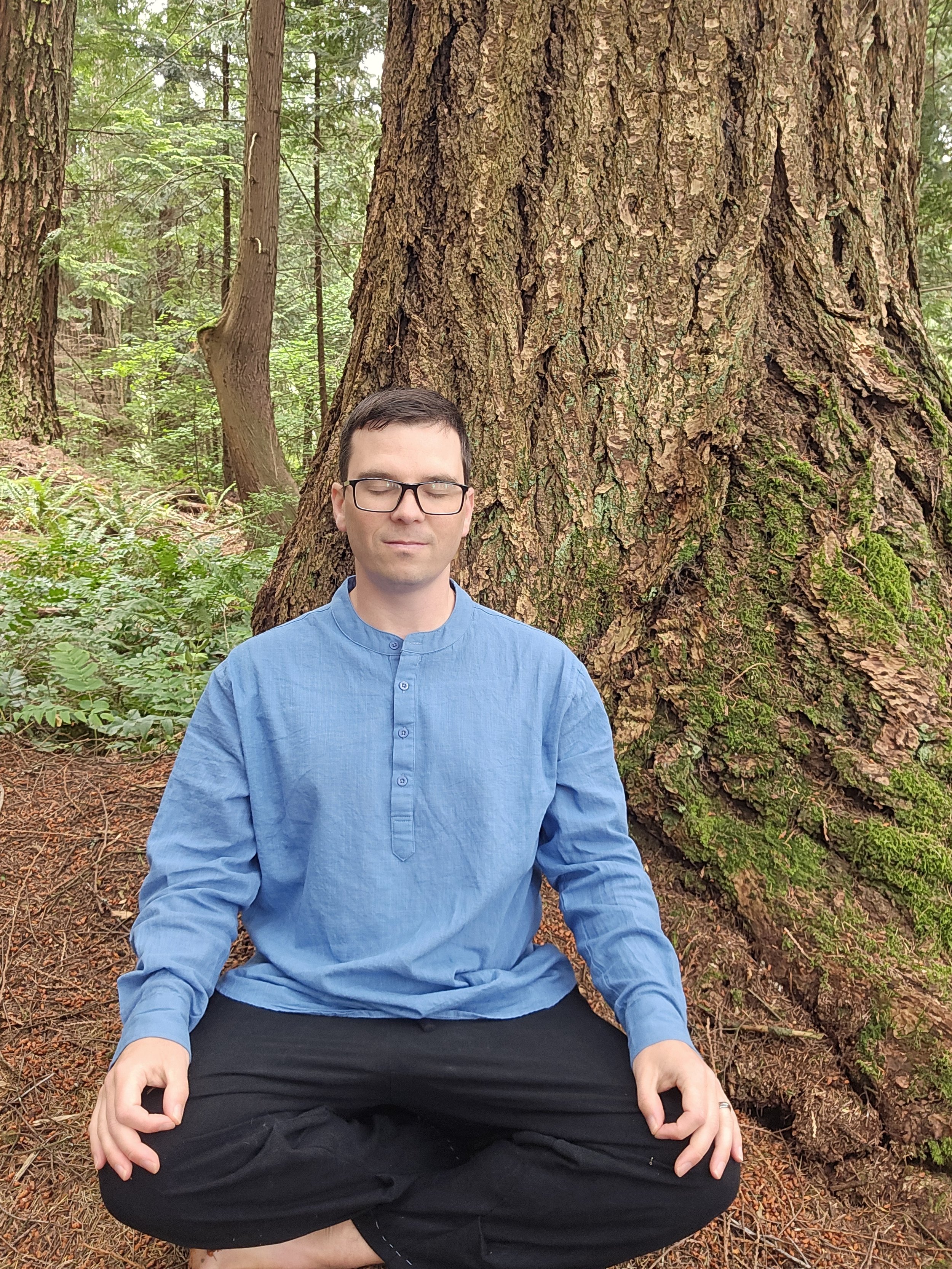 A man sitting cross-legged with eyes closed, meditating in front of a large moss-covered tree in a lush forest.