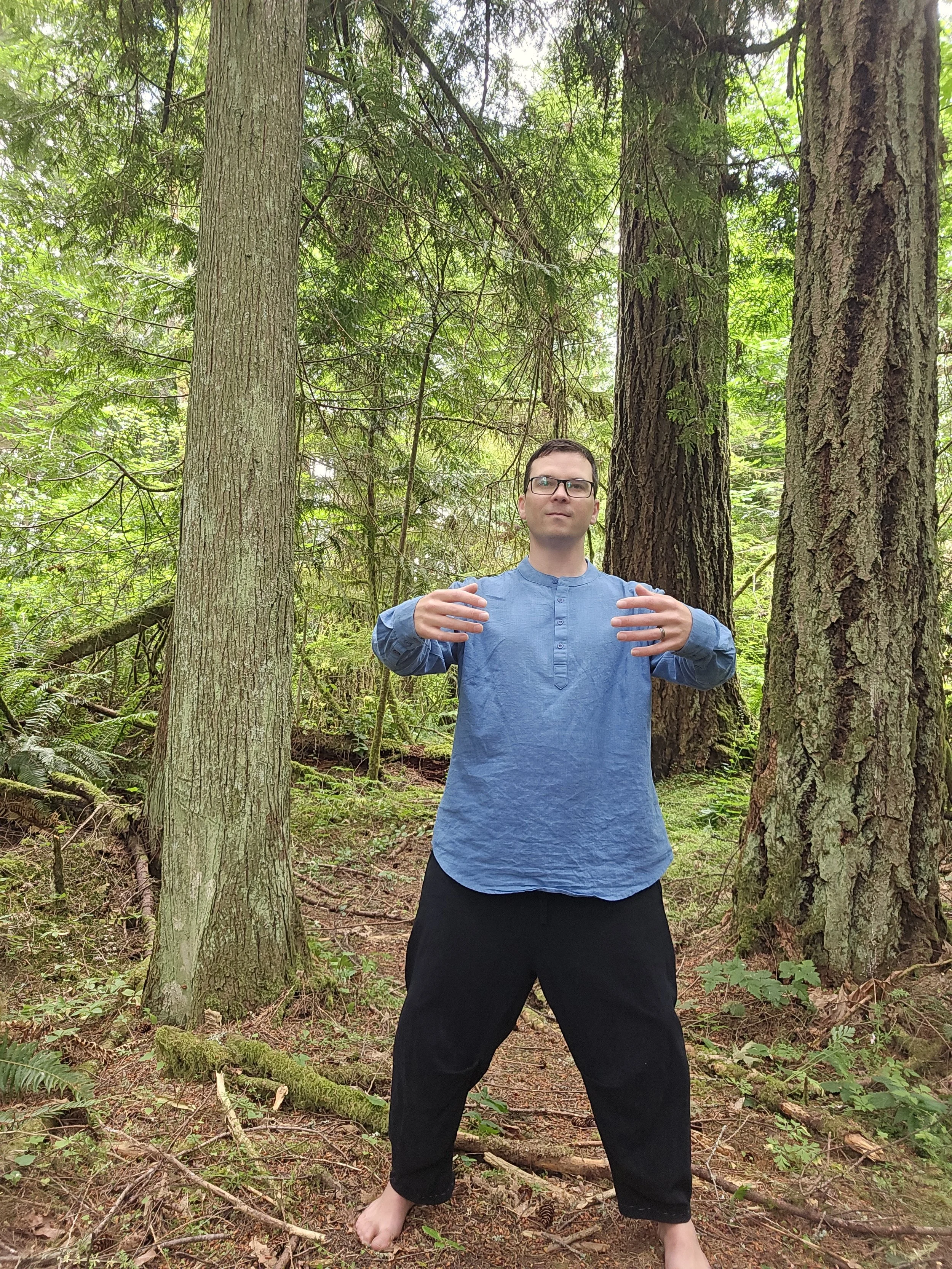 A man with glasses, wearing a blue long-sleeve shirt and black pants, stands barefoot in a forest with tall trees and green foliage, striking a meditative pose with arms raised and hands facing upward.