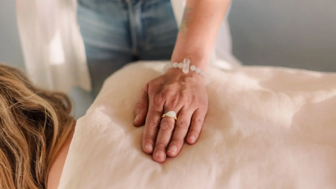 Carrie's hand is placed lightly on the back of a client who is lying comfortably face down on a massage table.