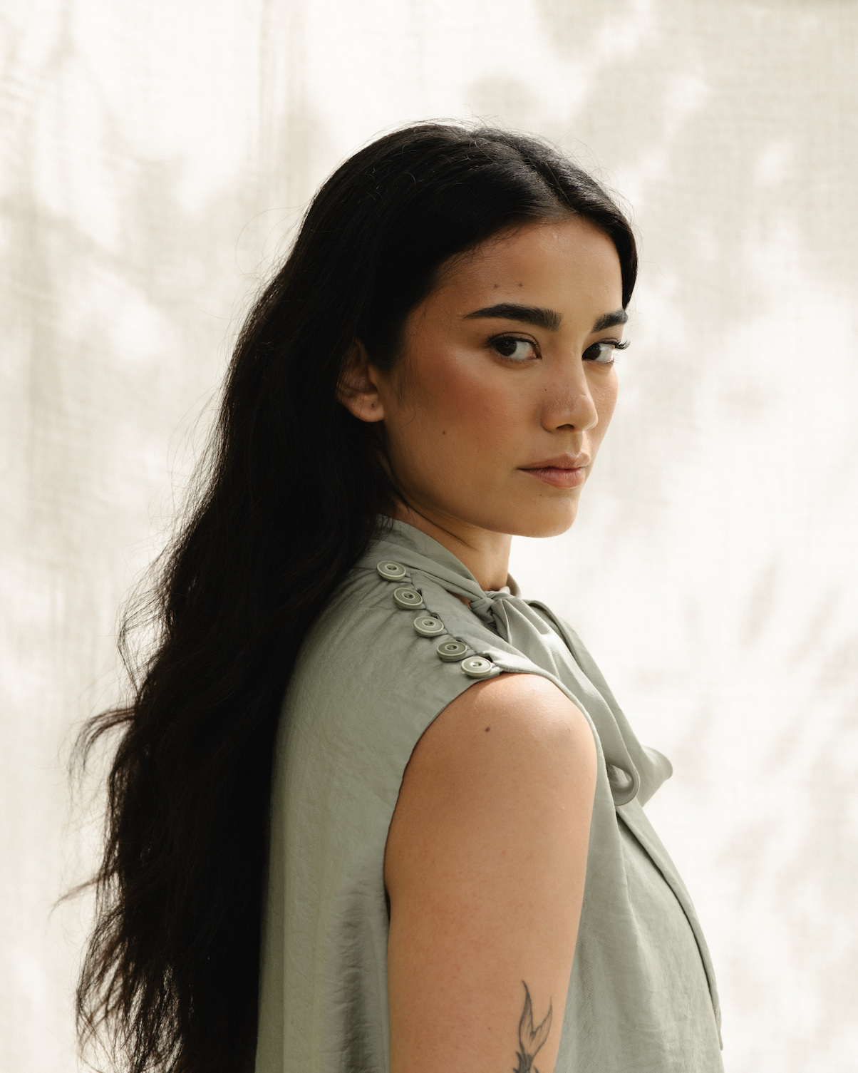 A woman with long dark hair and a tattoo on her upper arm, wearing a sleeveless gray top with button details on the shoulder, looking at the camera with a neutral expression in front of a light-colored curtain background.