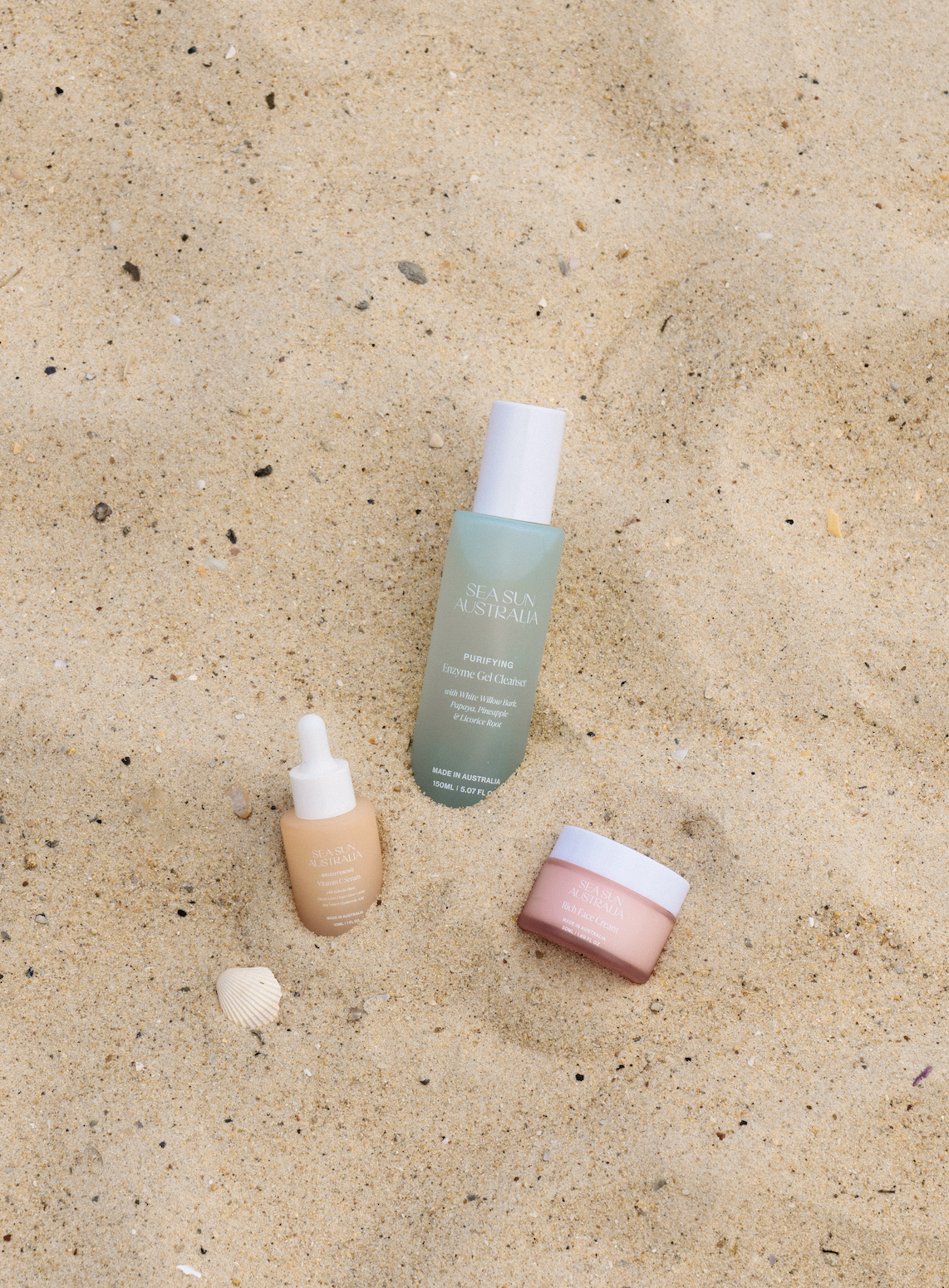 Skincare products on sandy beach, including a green Sea Sun Australia Purifying Enzyme Gel Cleanser, a beige Sea Sun Australia Vitamin C Serum with pipette, and a pink Sea Sun Australia Rich Face Cream in a jar.