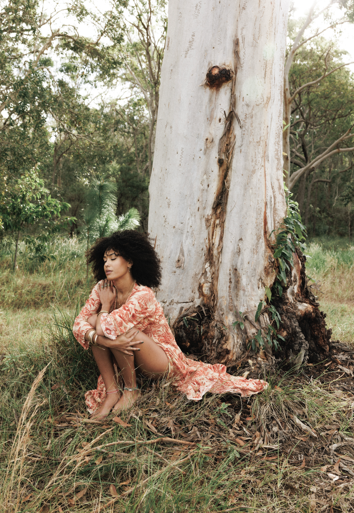 A woman with curly black hair in a floral dress sitting on the ground beside a large tree in a grassy forest area, with her eyes closed and arms crossed over her knees.