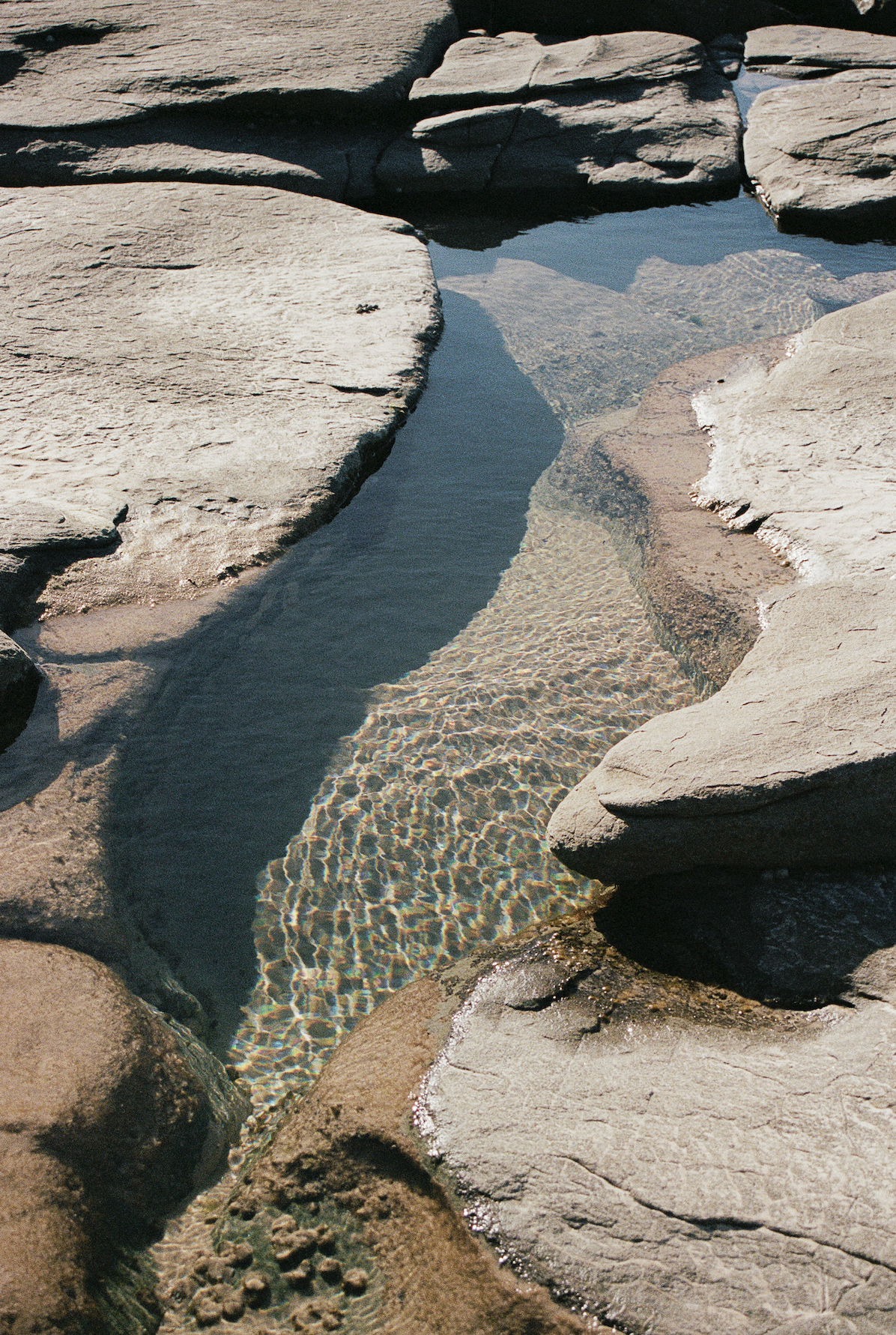 A narrow water channel with clear water flowing between large, flat rocks on a rocky shoreline.