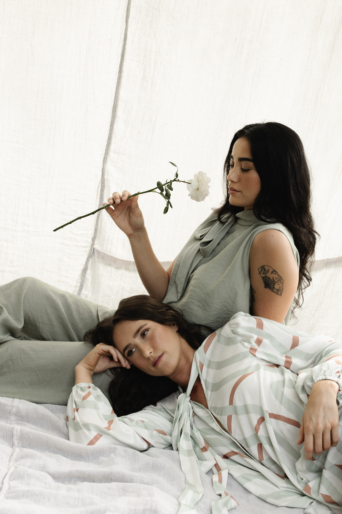 Two women relaxing on a bed in a light-colored tent. One woman is sitting and holding a white flower, while the other woman is lying down, resting her head on the first woman's lap. They are both dressed in soft, pastel-colored clothing.