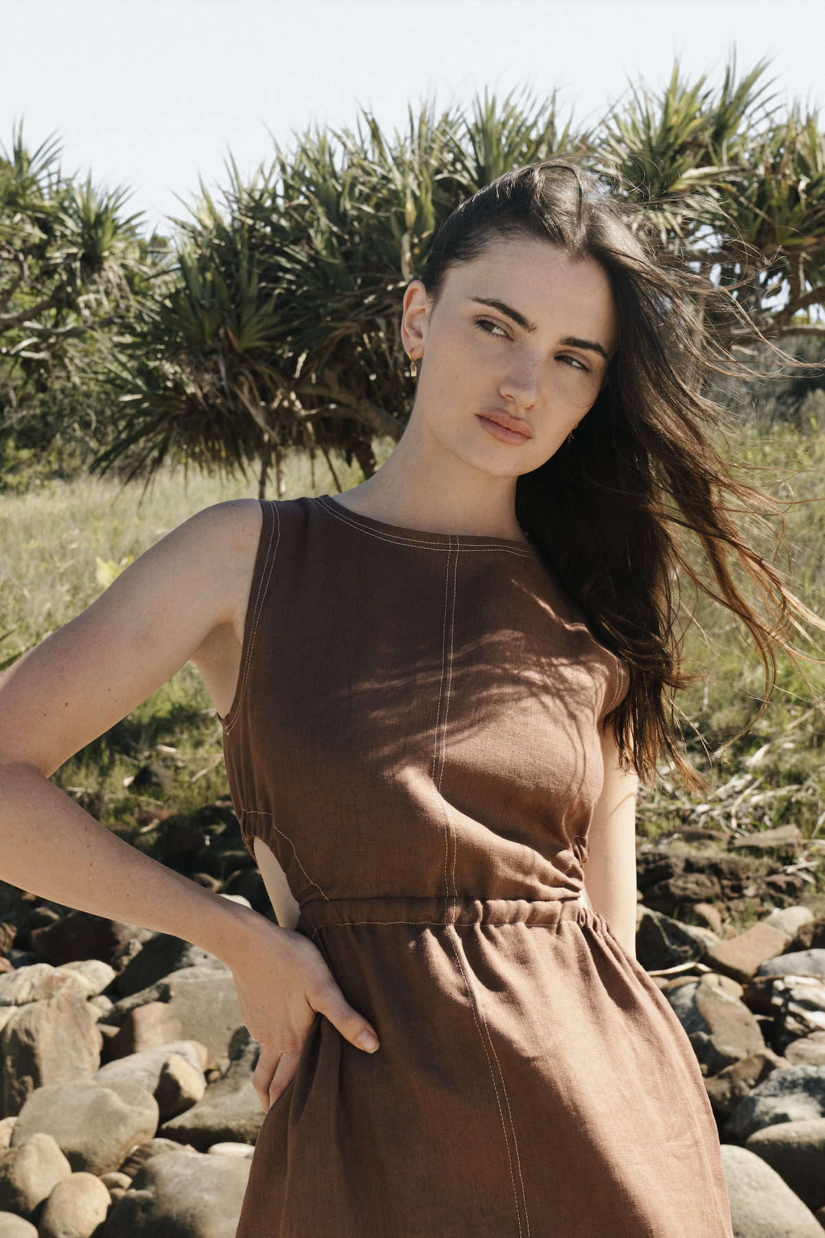A young woman with long brown hair, wearing a brown sleeveless dress, stands outdoors near rocks and greenery, looking into the distance during daytime.