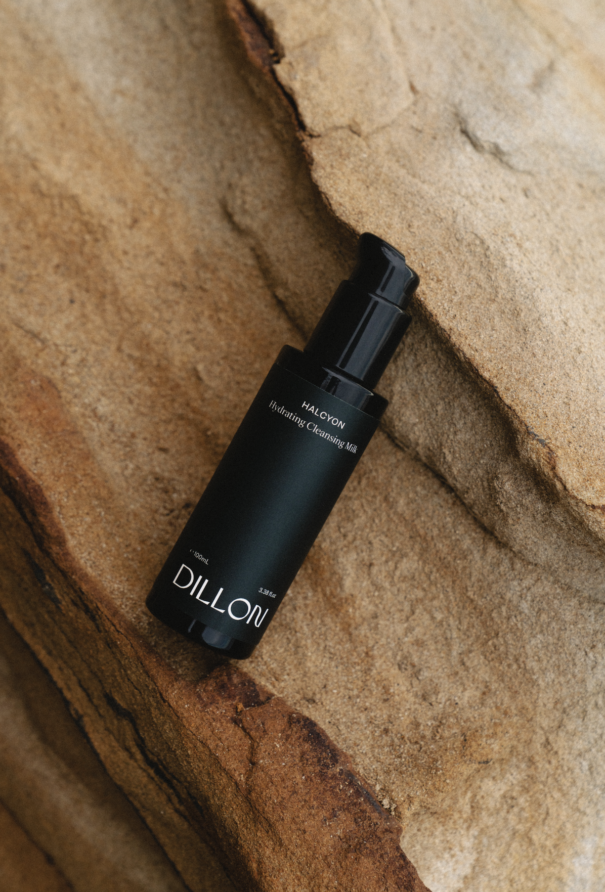 A black bottle of DILLON Hydrating Cleansing Milk lying on a rough, textured stone surface.