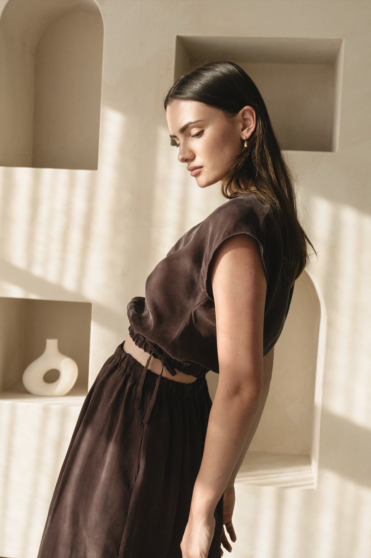 A woman with long dark hair in a brown satin outfit standing indoors with sunlight casting shadows on the wall behind her.