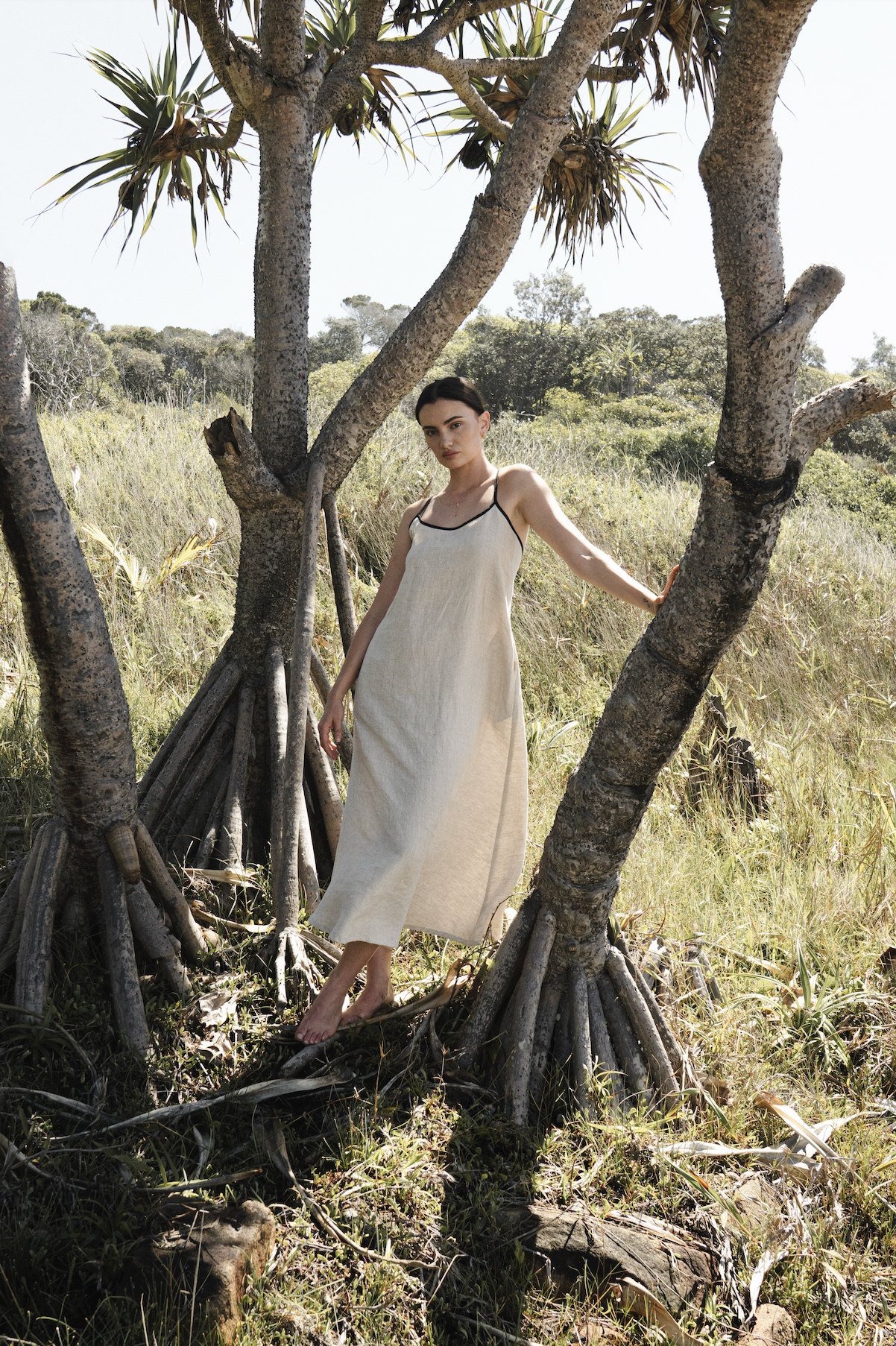 A woman in a light beige, sleeveless dress with black trim poses barefoot near a tree with exposed roots in a grassy, sunny field.