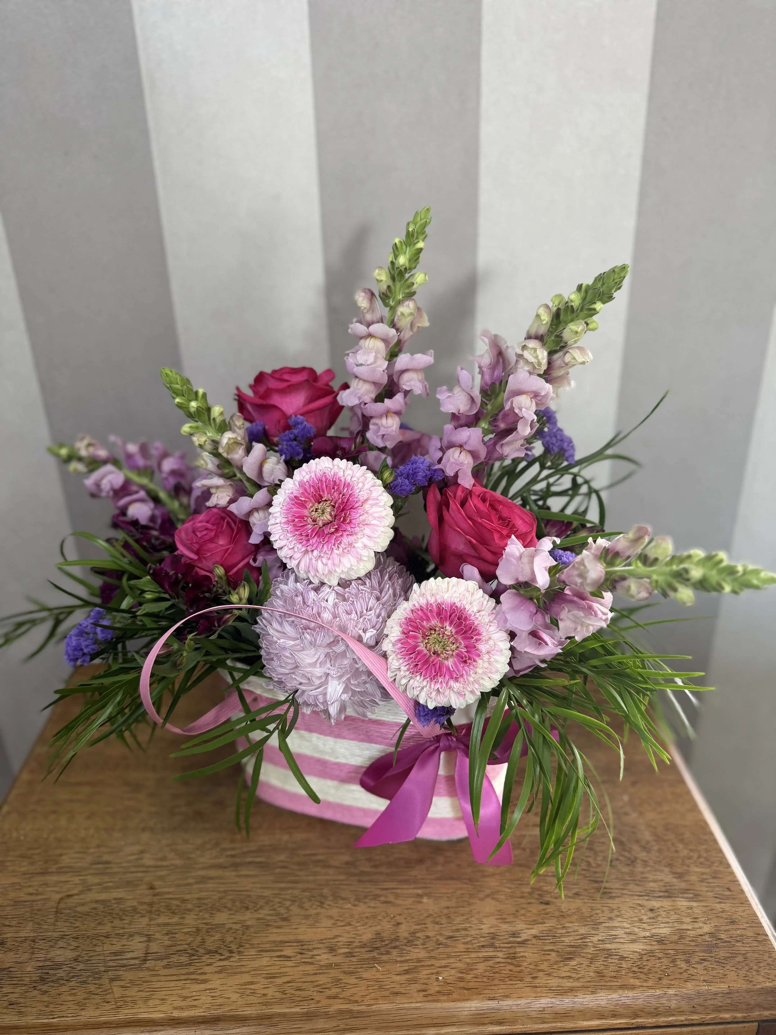 A colorful flower arrangement in a pink and white striped hatbox with a pink ribbon, featuring pink and purple flowers on a wooden surface.