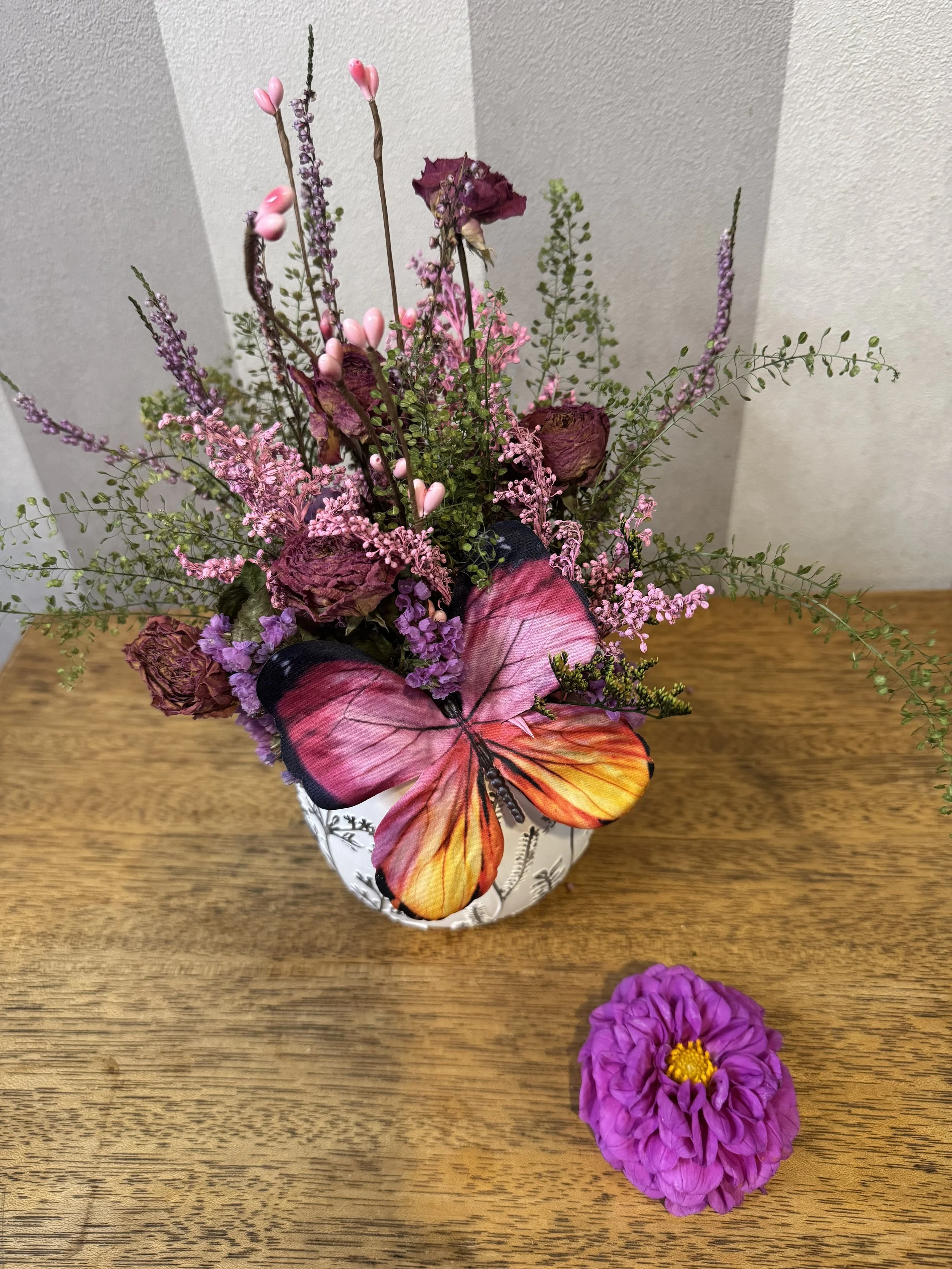 A flower arrangement with purple, pink, and dried flowers in a white vase on a wooden surface, and a purple flower lying next to the vase.