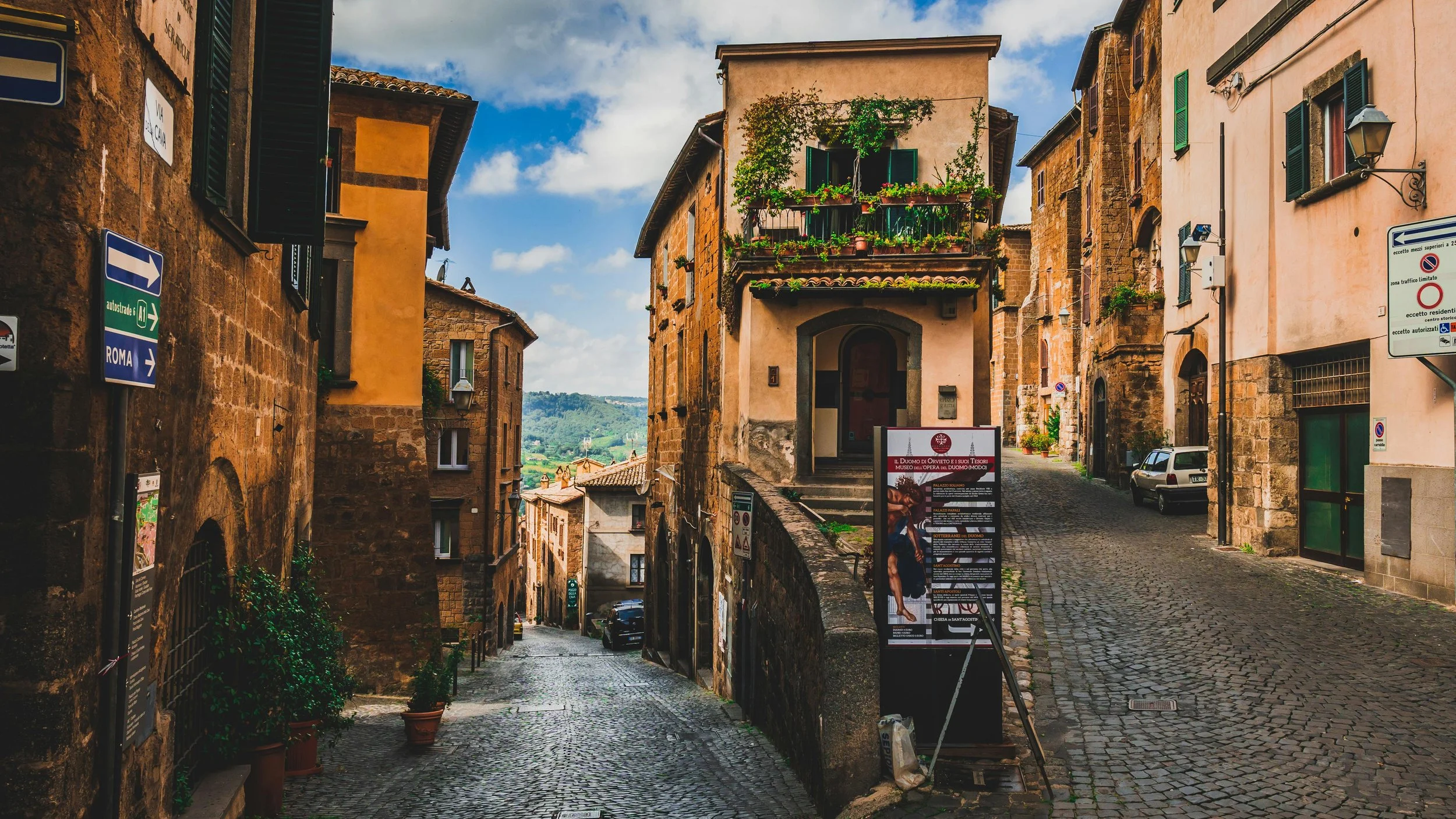 A narrow cobblestone street in a historic Italian hillside town with old stone buildings, some with flower-filled balconies, green shutters, and street signs, during daytime with partly cloudy skies.