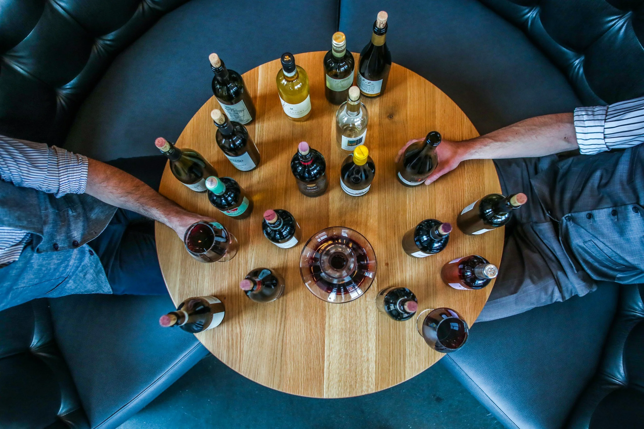 Overhead view of a round wooden table with various bottles of wine and a wine glass, surrounded by black leather seating.